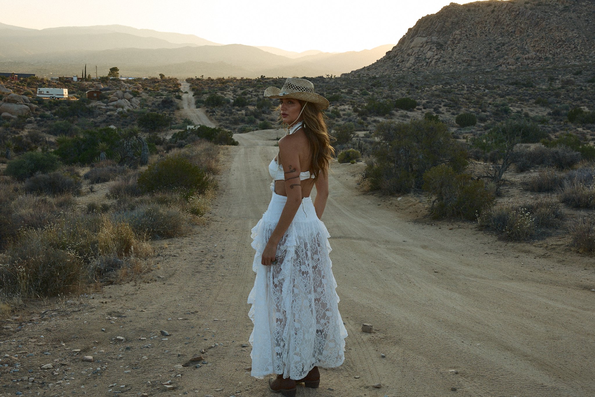 Woman in a white lace dress and straw hat standing on a dirt road in a desert landscape at sunset.