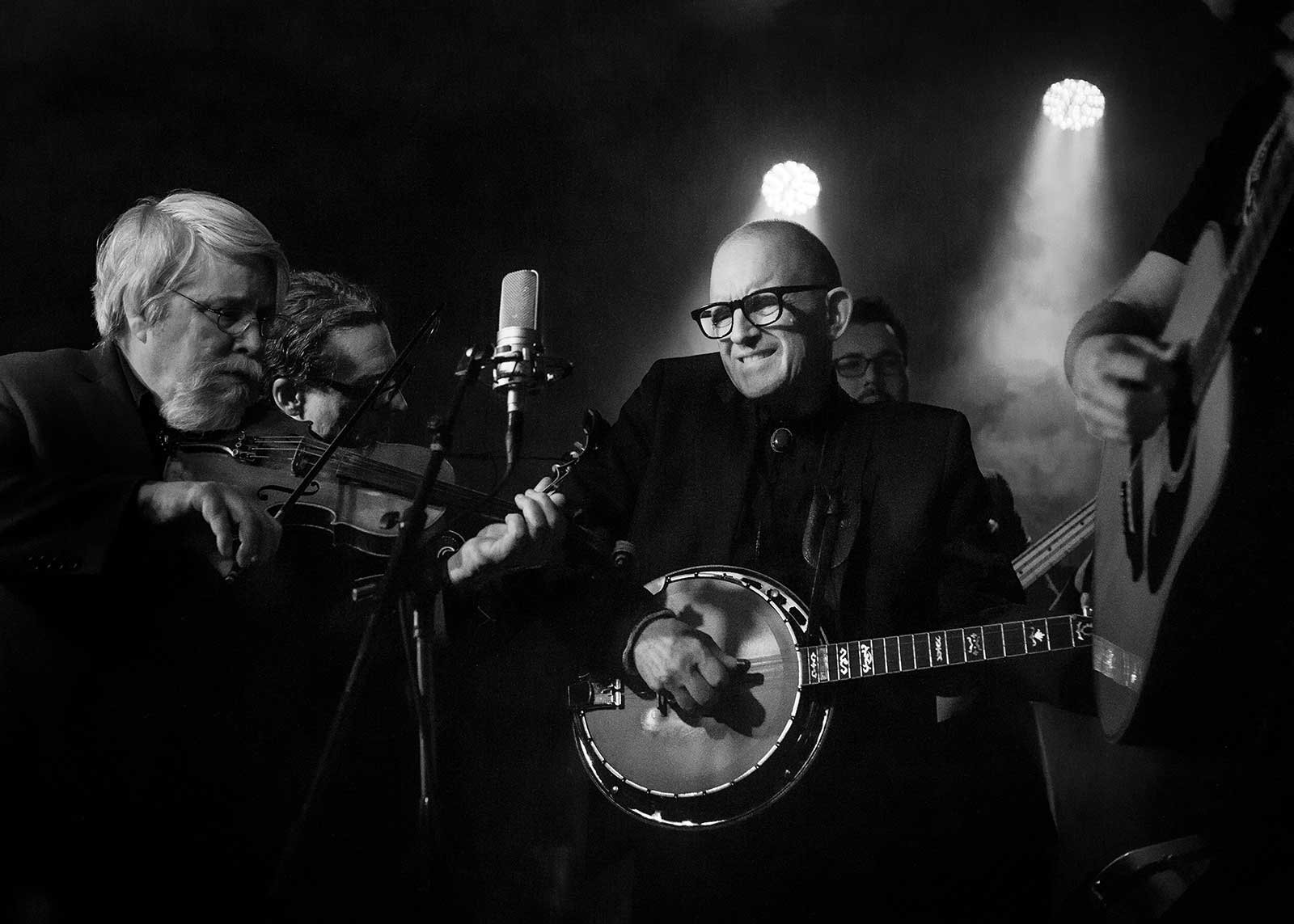 Musicians playing instruments on stage in a black and white photo, including a man with glasses and a banjo, a woman with a violin, and a person partially visible playing an acoustic guitar.