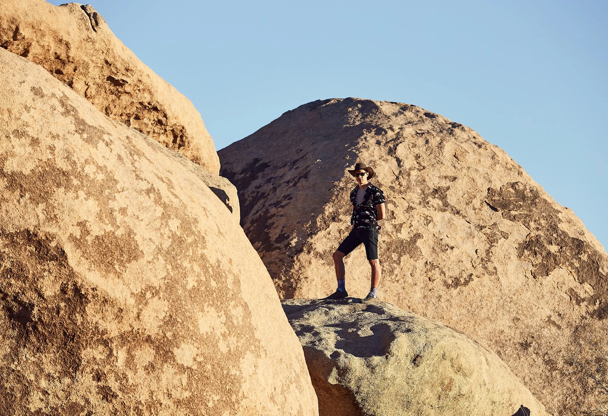 A person wearing a hat, sunglasses, and casual clothes standing on a large rock formation in a desert landscape with big boulders and a clear blue sky.