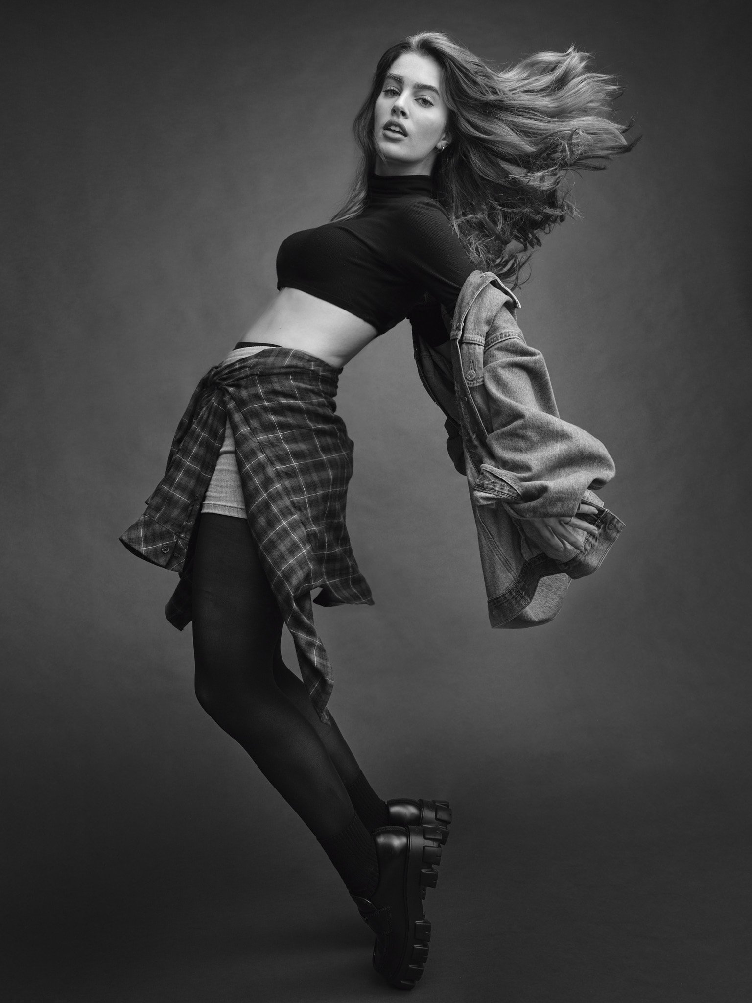 Black and white photo of a young woman with flowing hair, wearing a crop top, jacket, plaid shirt, tights, and boots, posing dynamically against a plain background.