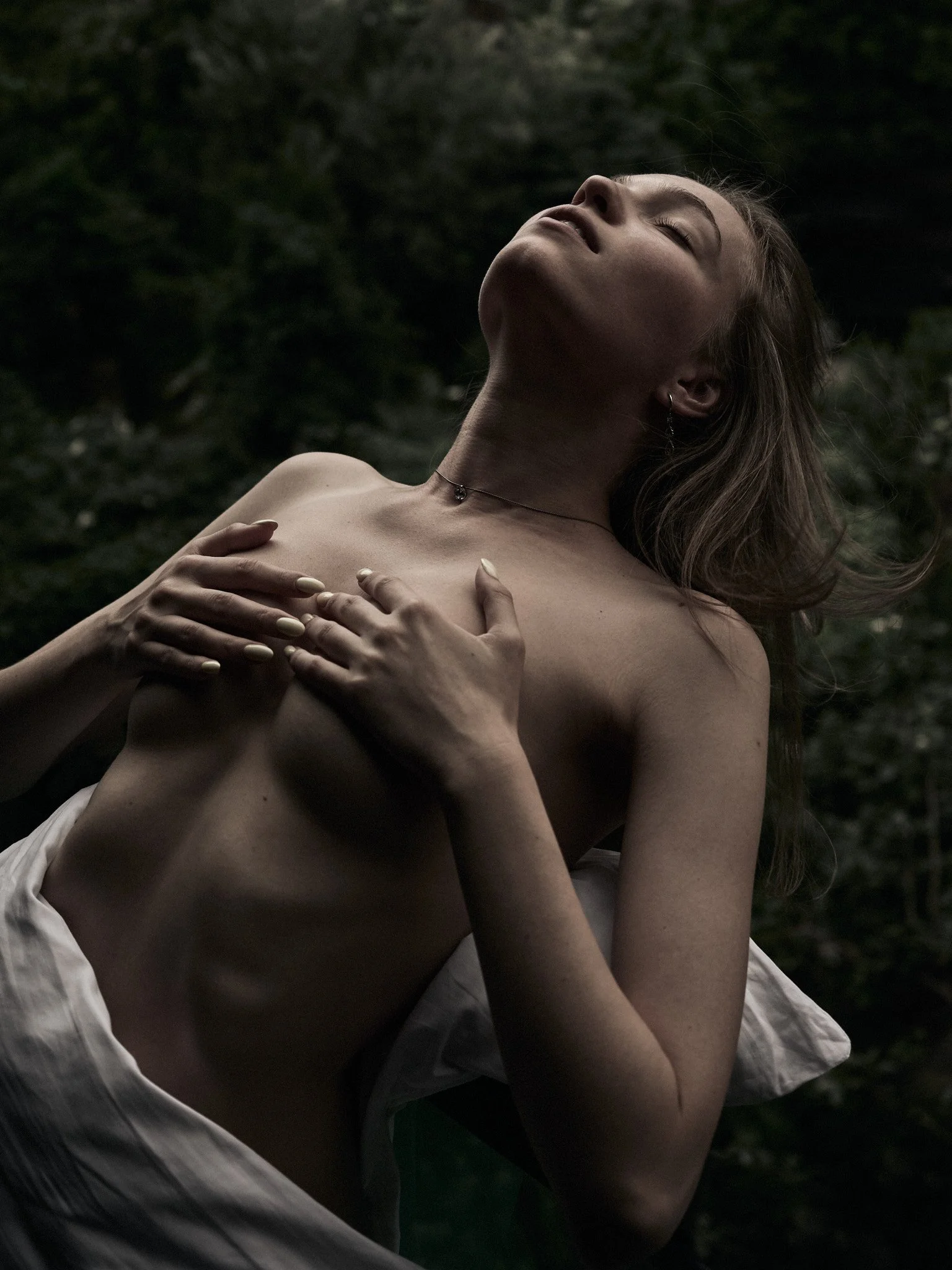 A woman with long hair and fair skin poses outdoors, using her hands to cover her chest, with her head tilted back and eyes closed, with a lush green background.