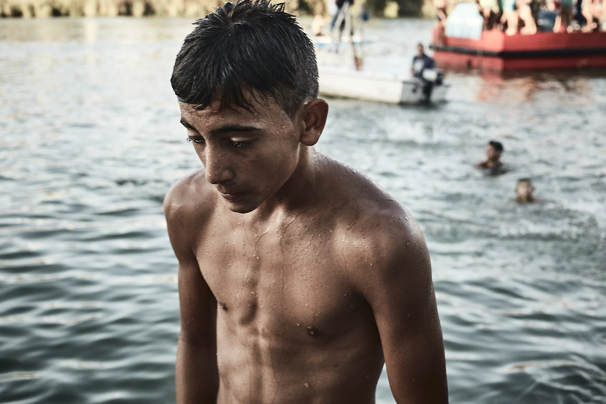 A young man with wet hair and a bare torso standing in a body of water with people swimming and boats in the background.