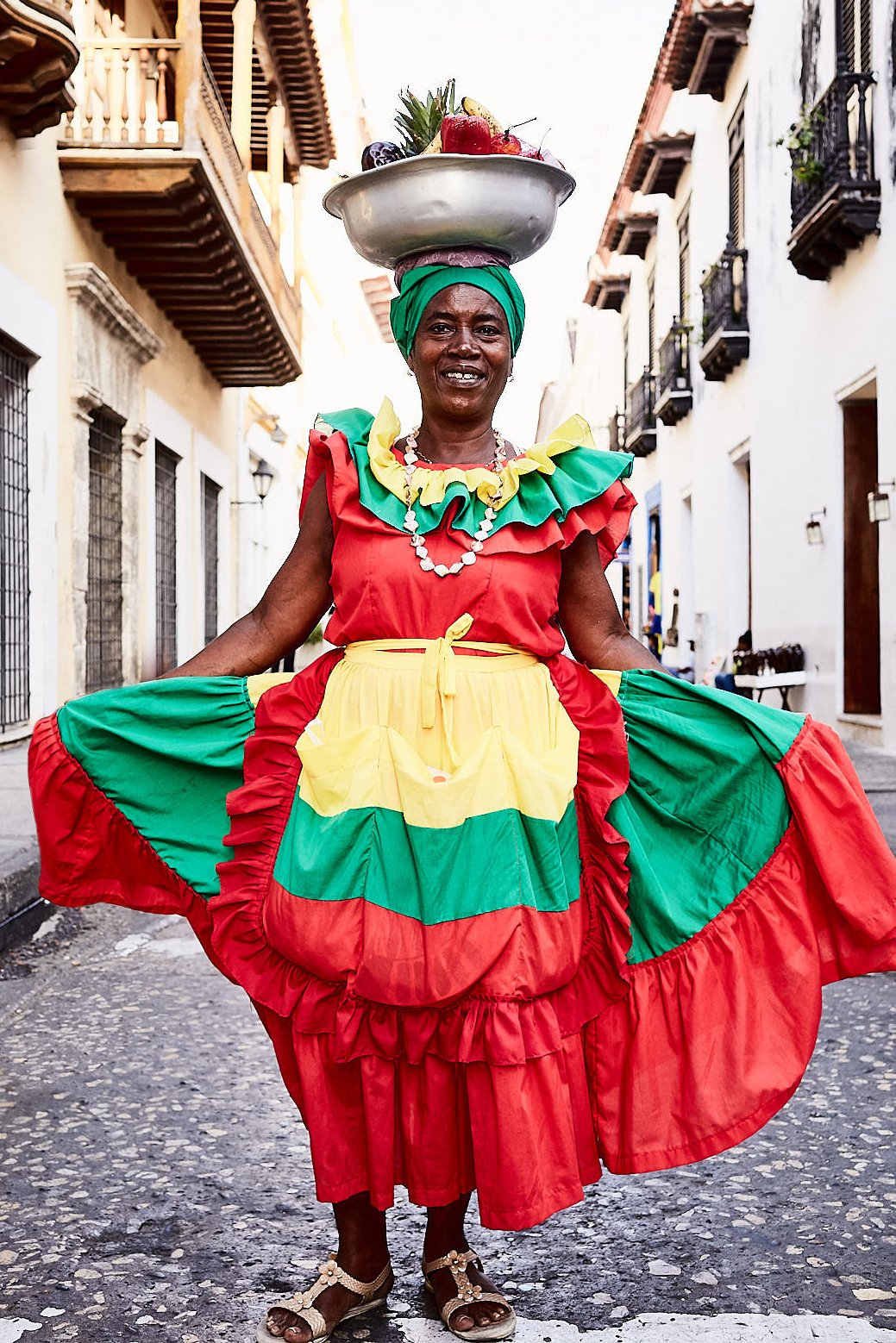 An older woman dressed in a colorful red, yellow, green, and black traditional dress, standing on a cobblestone street with white buildings around her. She is balancing a large bowl of fruits on her head and holding out her dress, smiling at the came