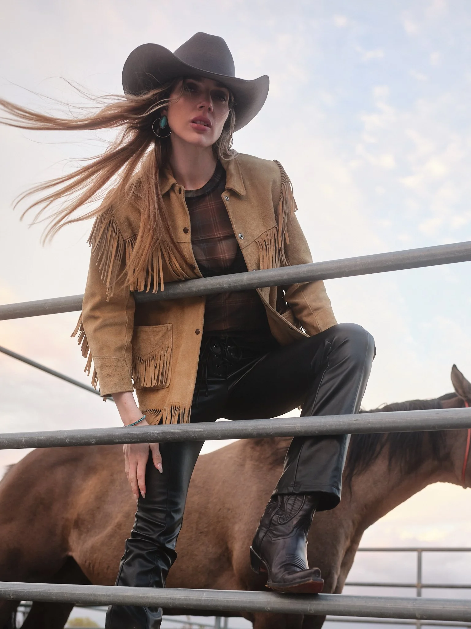 A woman wearing a cowboy hat, fringe leather jacket, plaid shirt, and black leather pants, sitting on a fence with a horse in the background during a cloudy day.