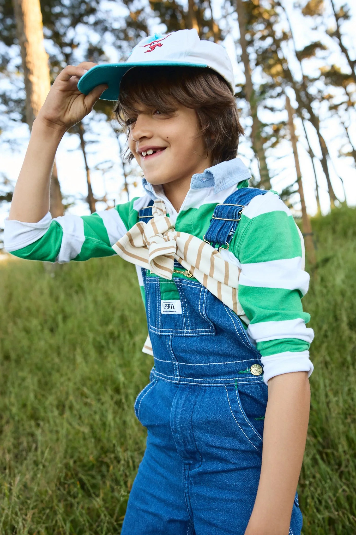 Young boy outdoors in a forest, wearing a baseball cap, striped shirt, striped scarf, and denim overalls, smiling and holding the brim of his cap.