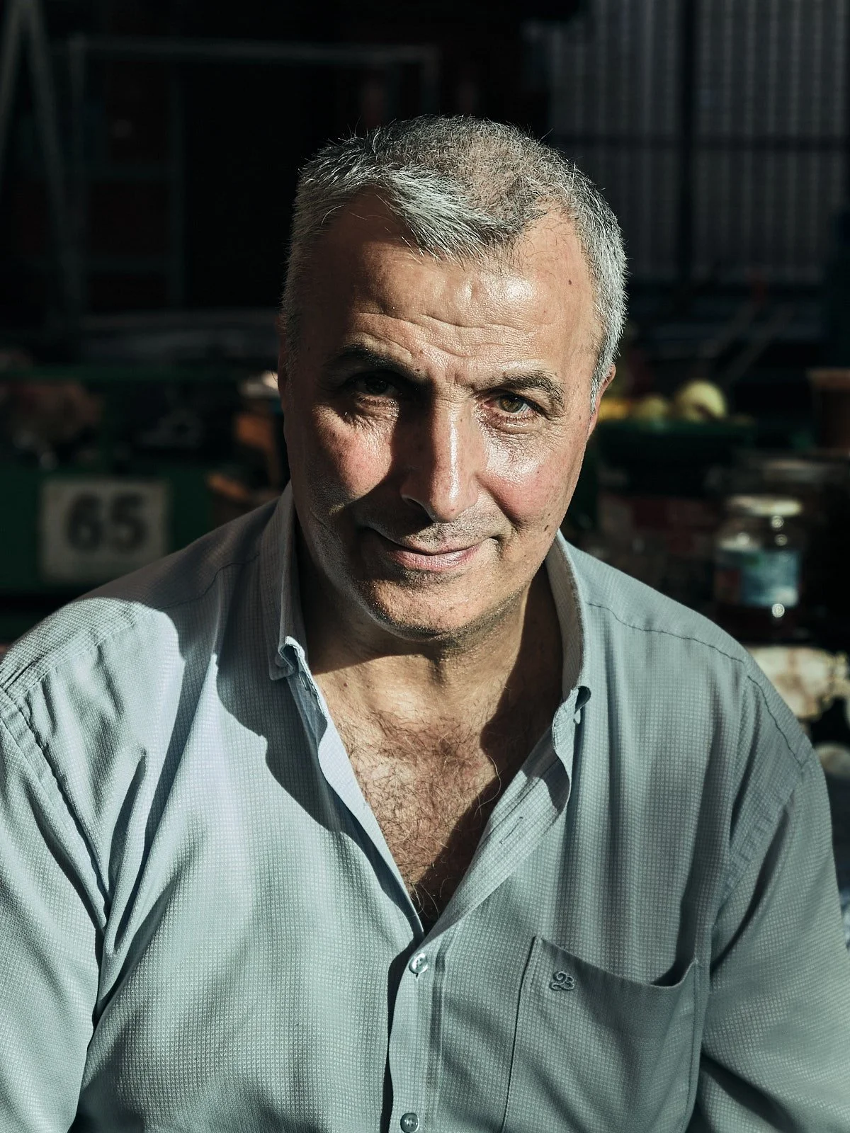 Close-up photo of a middle-aged man with short gray hair, wearing a light gray collared shirt, outdoors in sunlight, with a blurred background of market stalls or outdoor market.