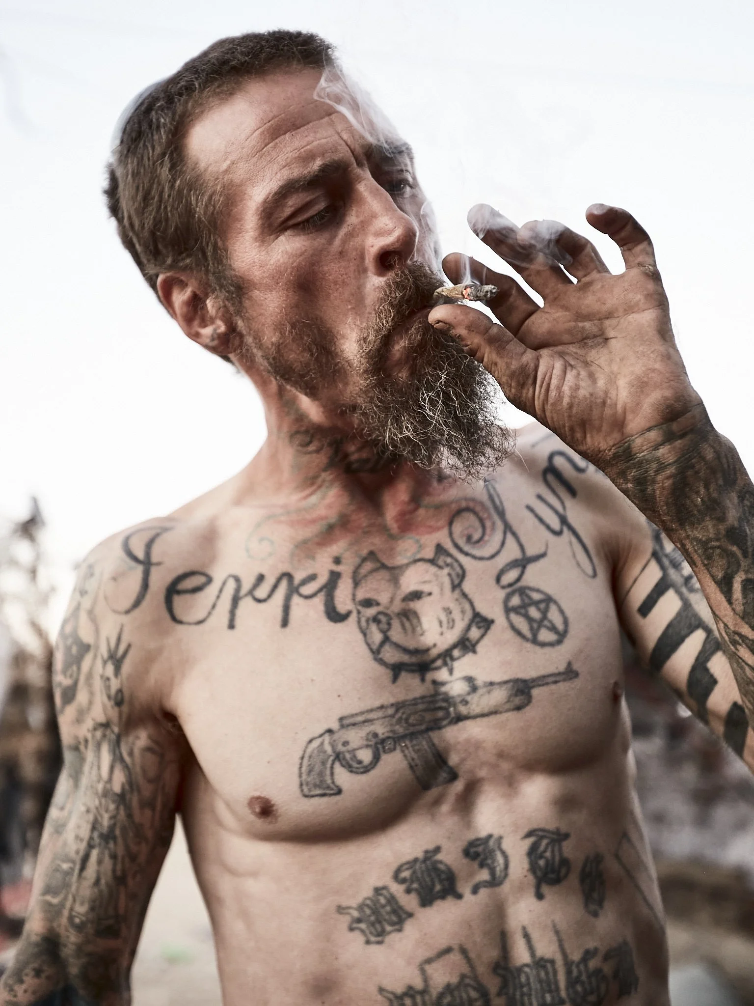 A shirtless man with a beard and tattoos on his chest and arms is smoking a cigarette outdoors.