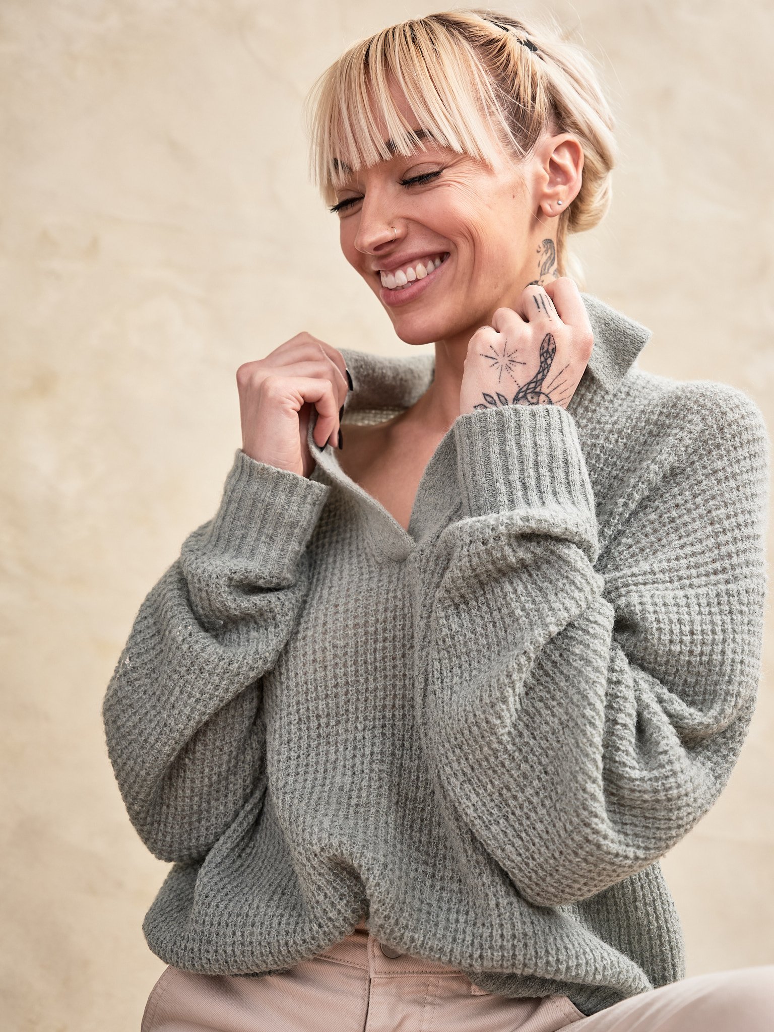 A woman with blonde hair and tattoos smiling while wearing a gray knit sweater and light-colored pants, standing against a beige background.