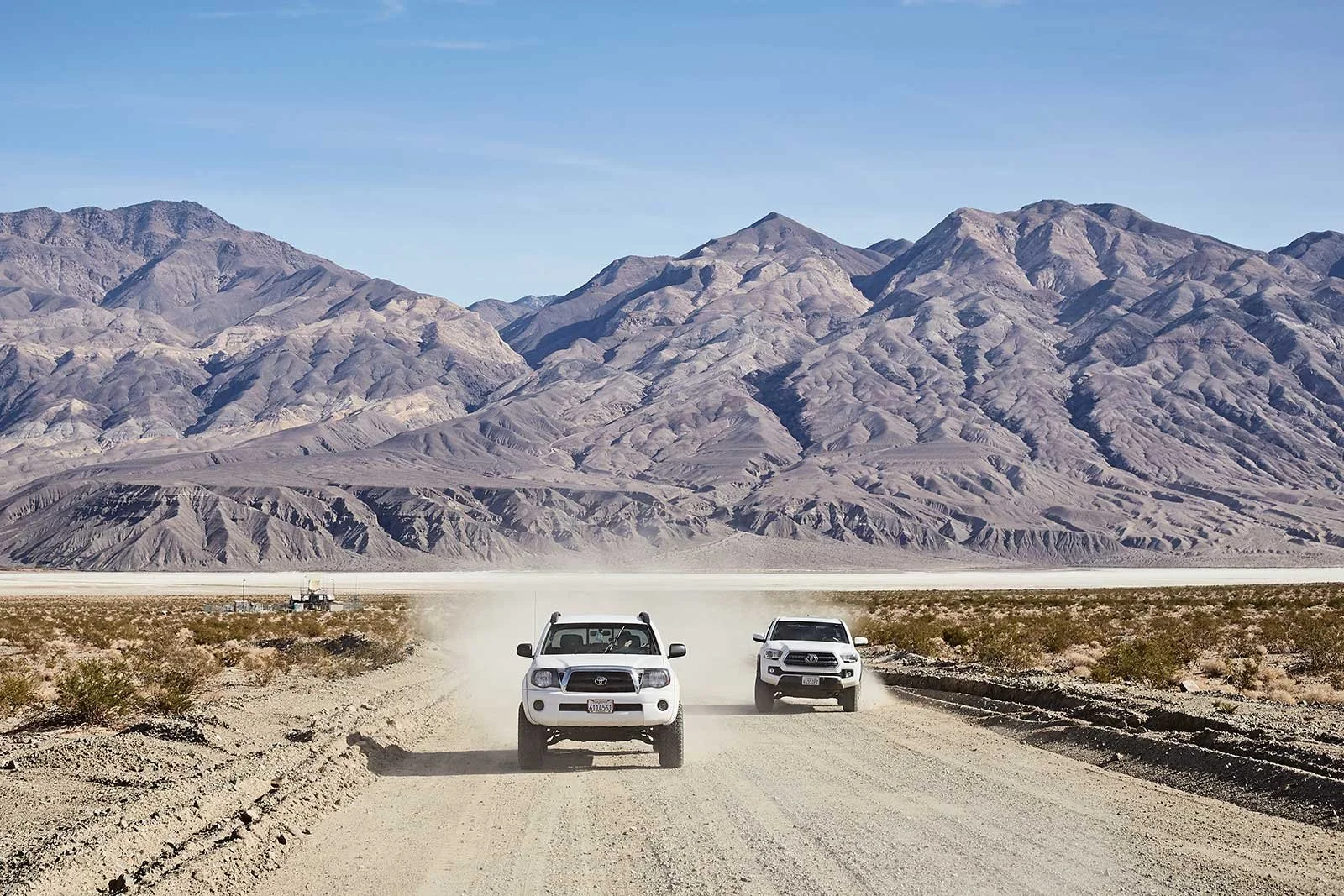 Two white pickup trucks driving on a dusty desert road with mountains in the background.