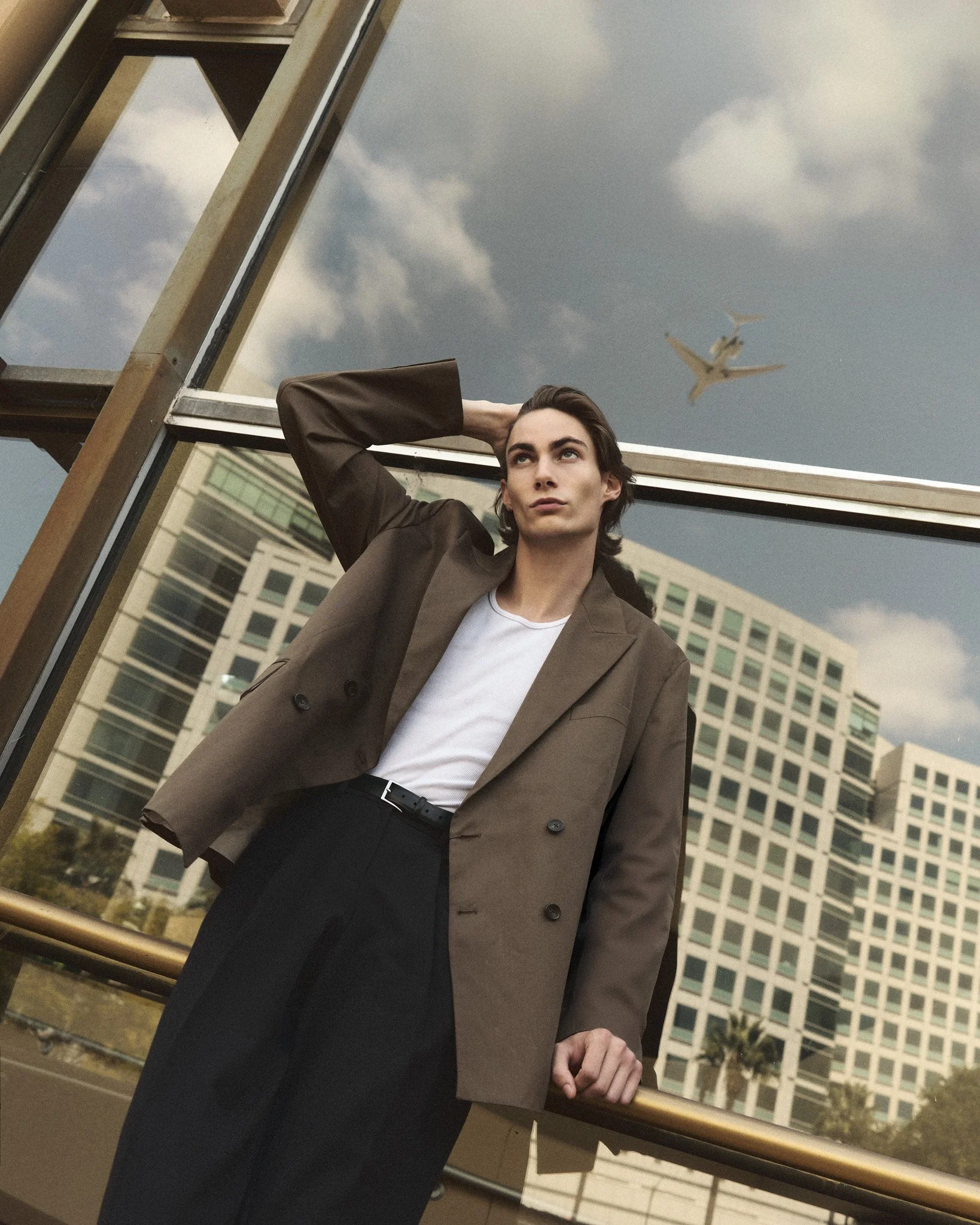A man wearing a brown blazer and dark slacks standing outside, leaning on a railing with a cityscape and an airplane in the sky behind him.