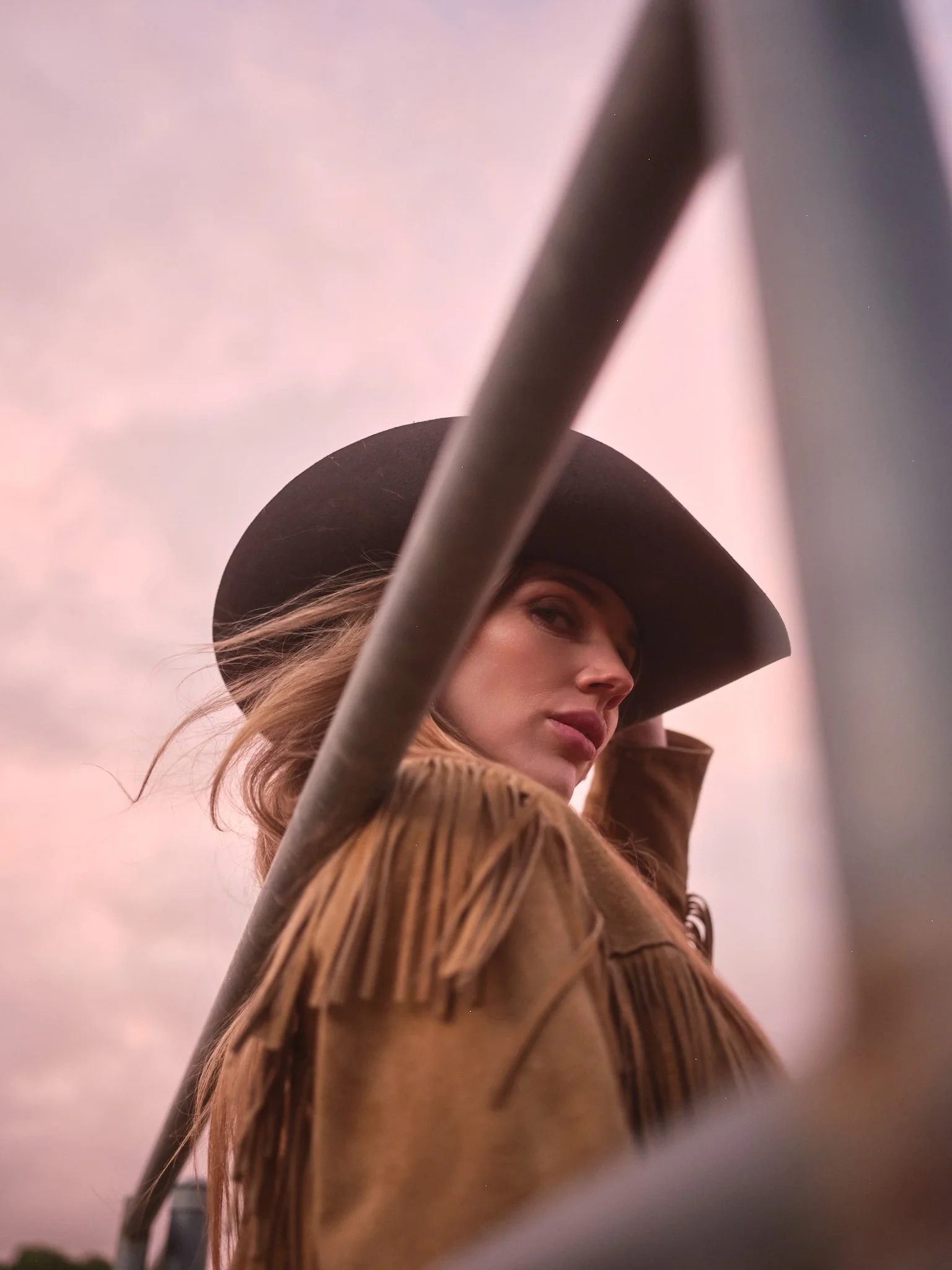 A woman wearing a wide-brimmed hat and fringed jacket looking over her shoulder with a pinkish sky in the background.