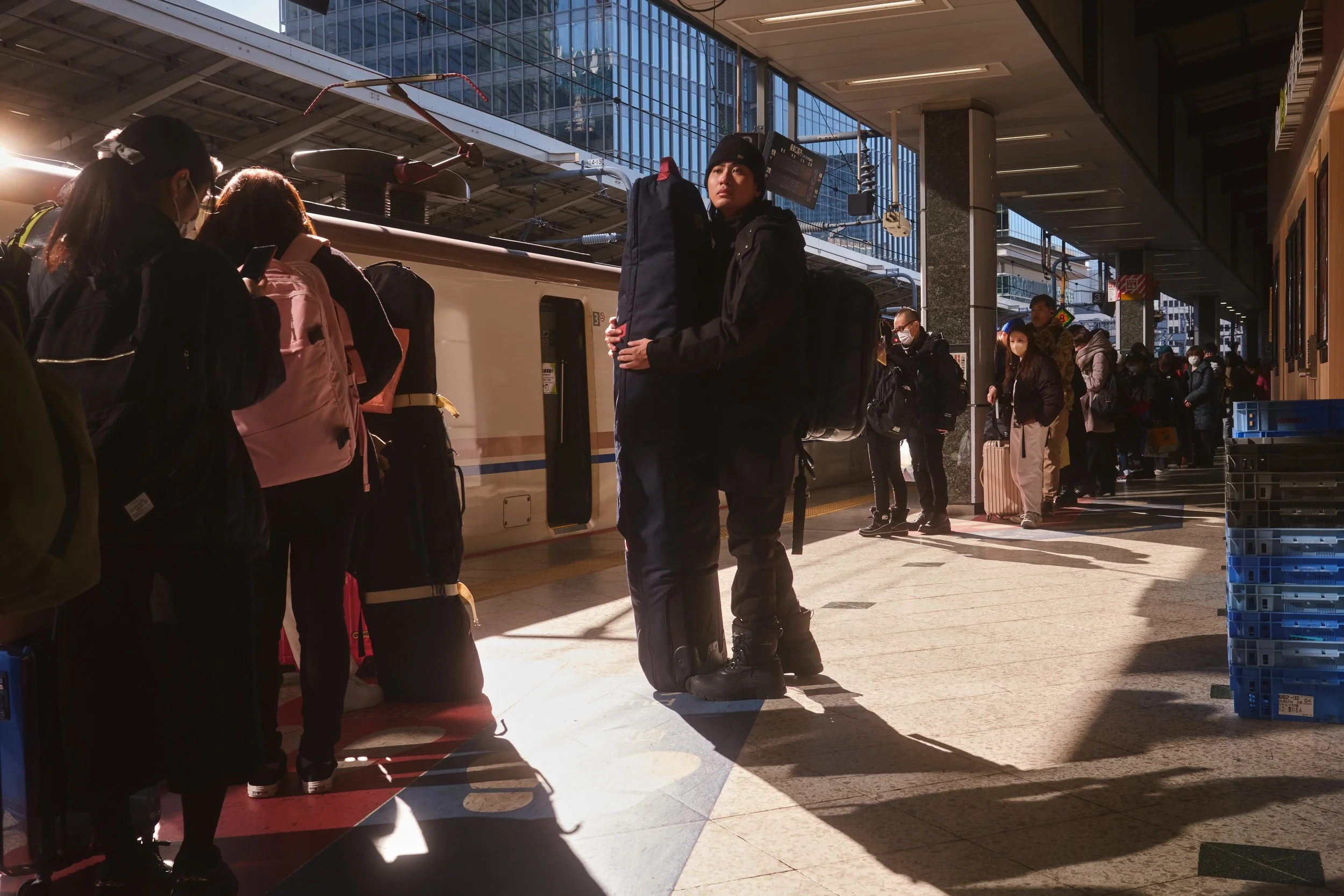 People standing in line at a train station platform, some with luggage, with a train in the background and sunlight casting shadows on the ground.