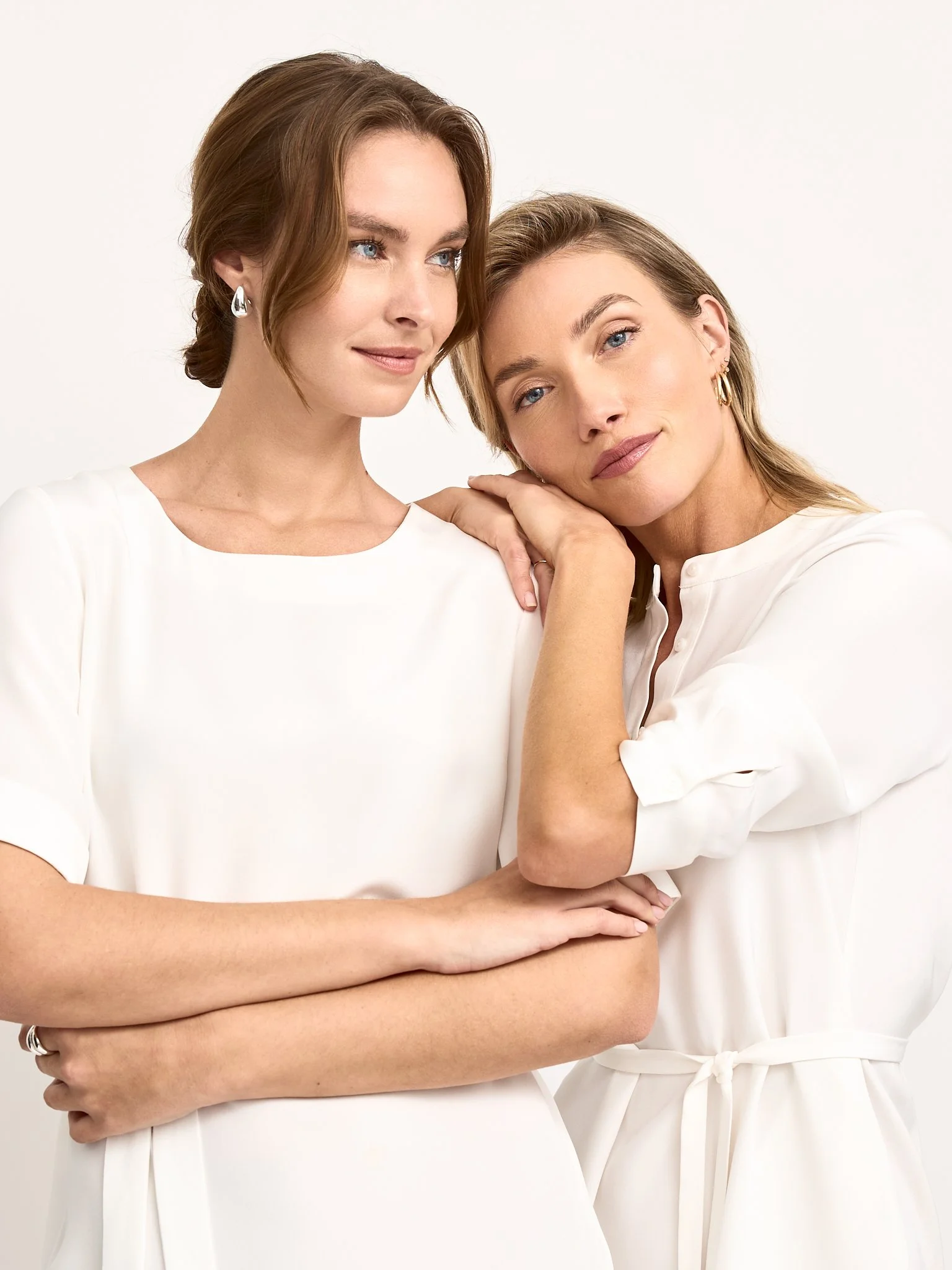 Two women dressed in white dresses standing close together, with one gently resting her head on the other's shoulder, against a plain light background.