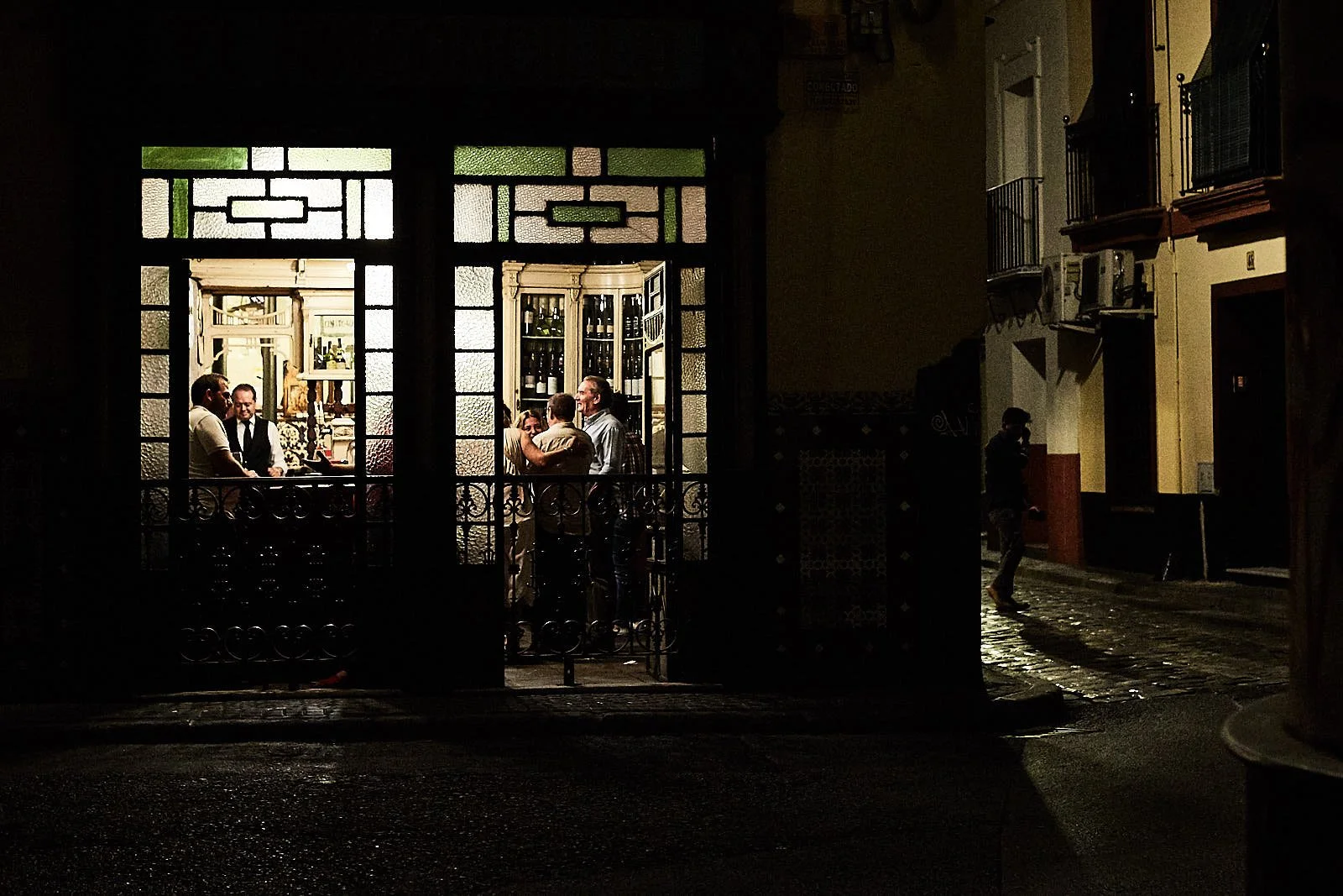 A nighttime scene of a sidewalk with a lit restaurant or bar visible through a window, showing people inside engaging in conversation.