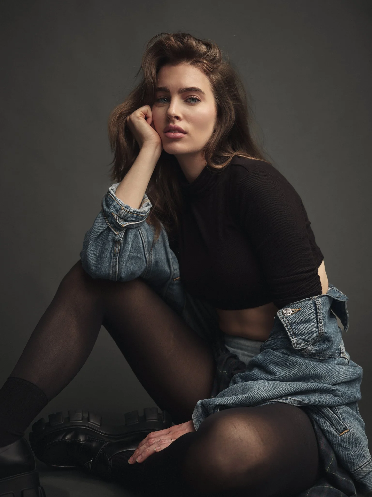 A young woman with wavy brown hair poses sitting on the floor against a dark gray background. She is wearing a black crop top, denim shorts, black stockings, and chunky black boots. One hand is resting on her knee while her head rests on her other ha