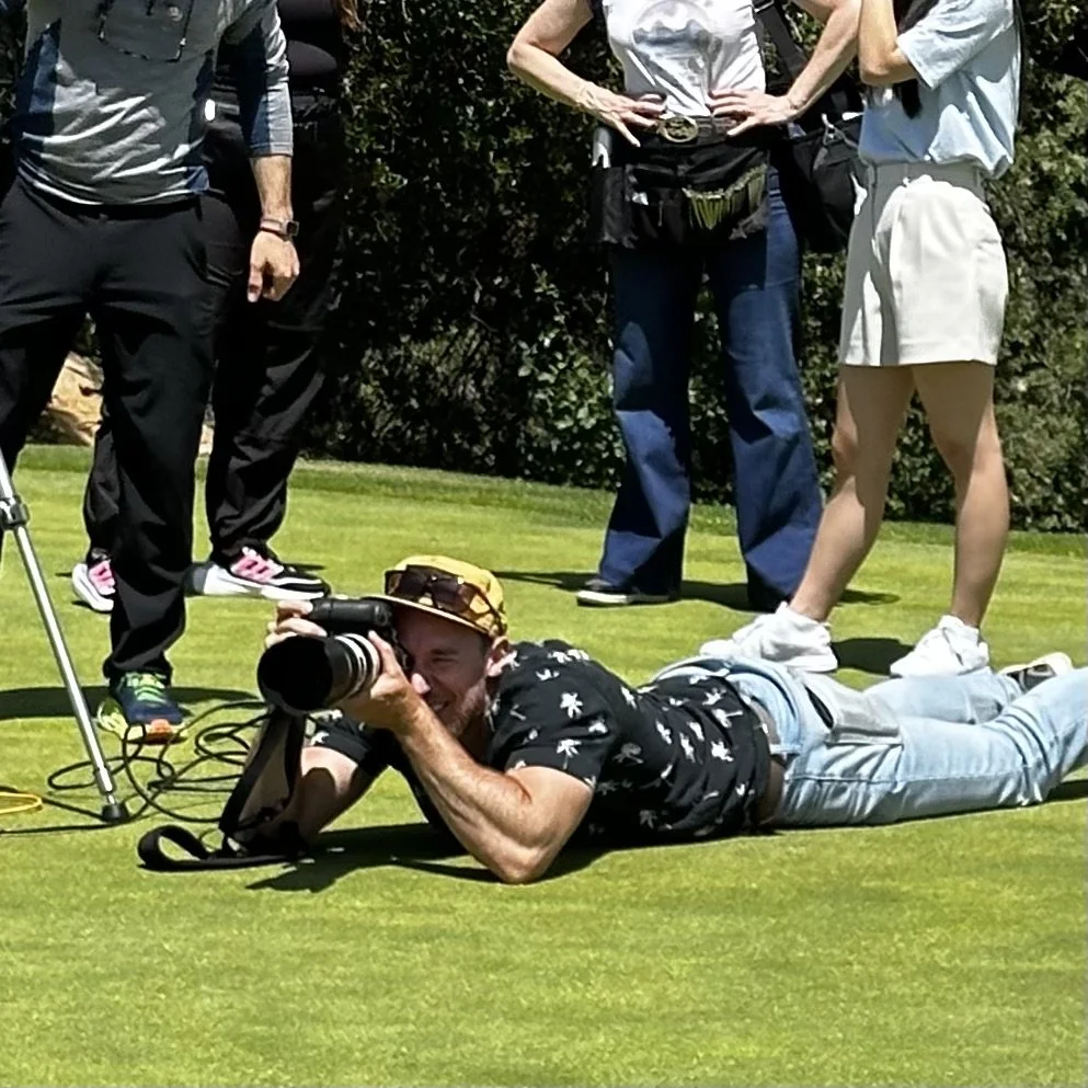 A man with a camera lying on the grass, taking photos at an outdoor event, with several people standing nearby.