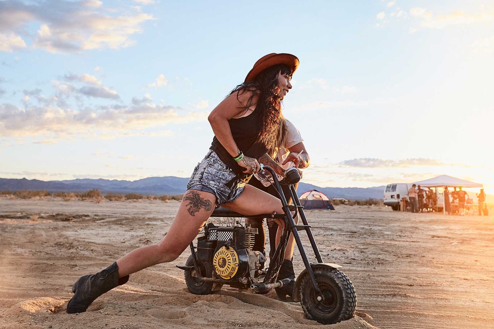 A woman with long hair, wearing a cowboy hat, shorts, and boots, riding a small mini bike on sandy terrain during sunset. In the background, there are tents, a van, and a group of people near a canopy.