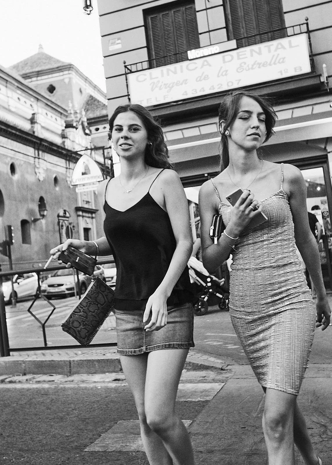Two young women walk on a city sidewalk. One wears a spaghetti strap top and jean skirt, the other a striped dress and holds a phone. Buildings and cars are in the background.
