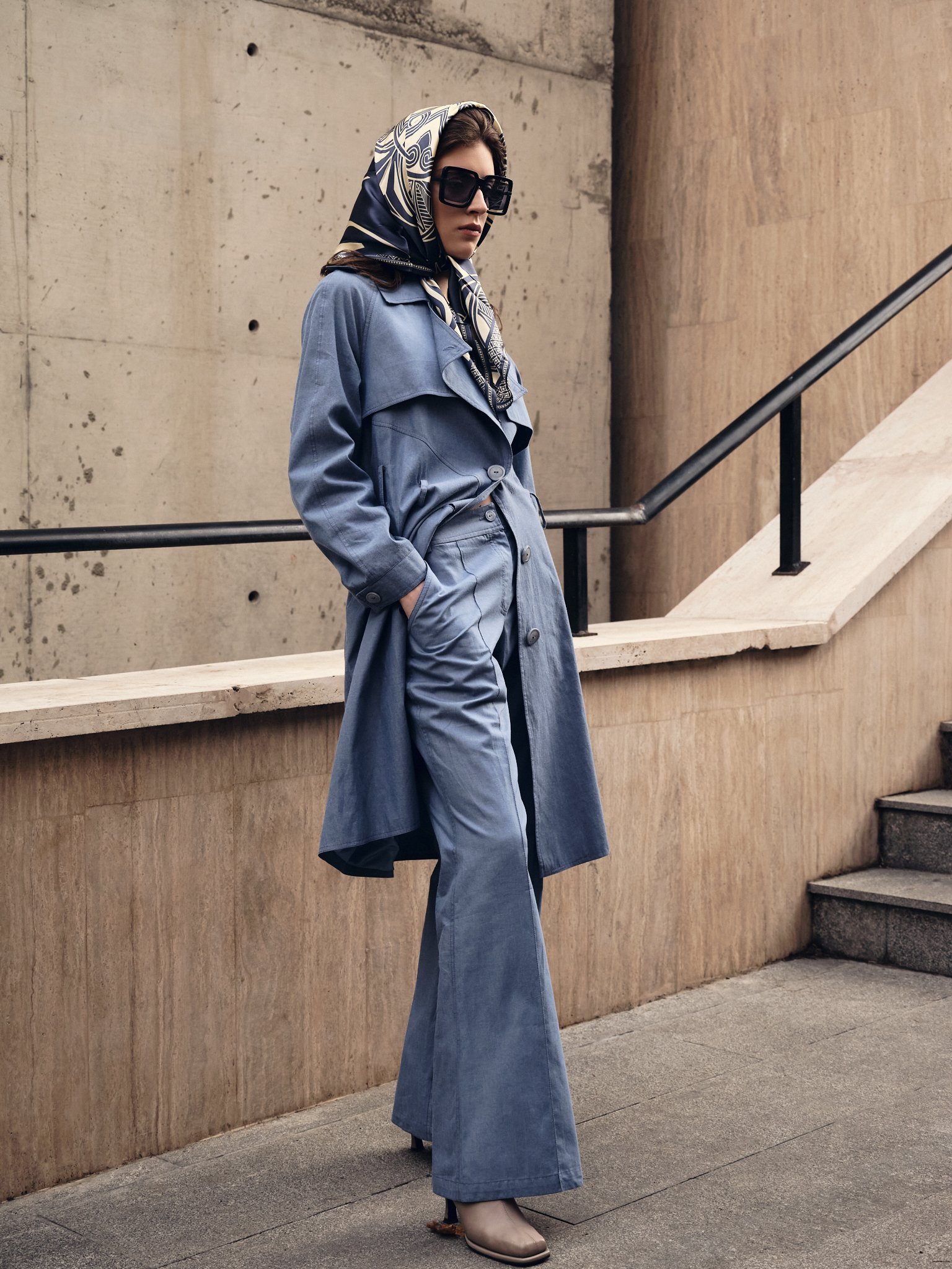 A woman with dark hair, wearing a blue trench coat, matching wide-leg jeans, large black sunglasses, a patterned headscarf, and beige ankle boots, standing on an outdoor staircase with a concrete wall background.