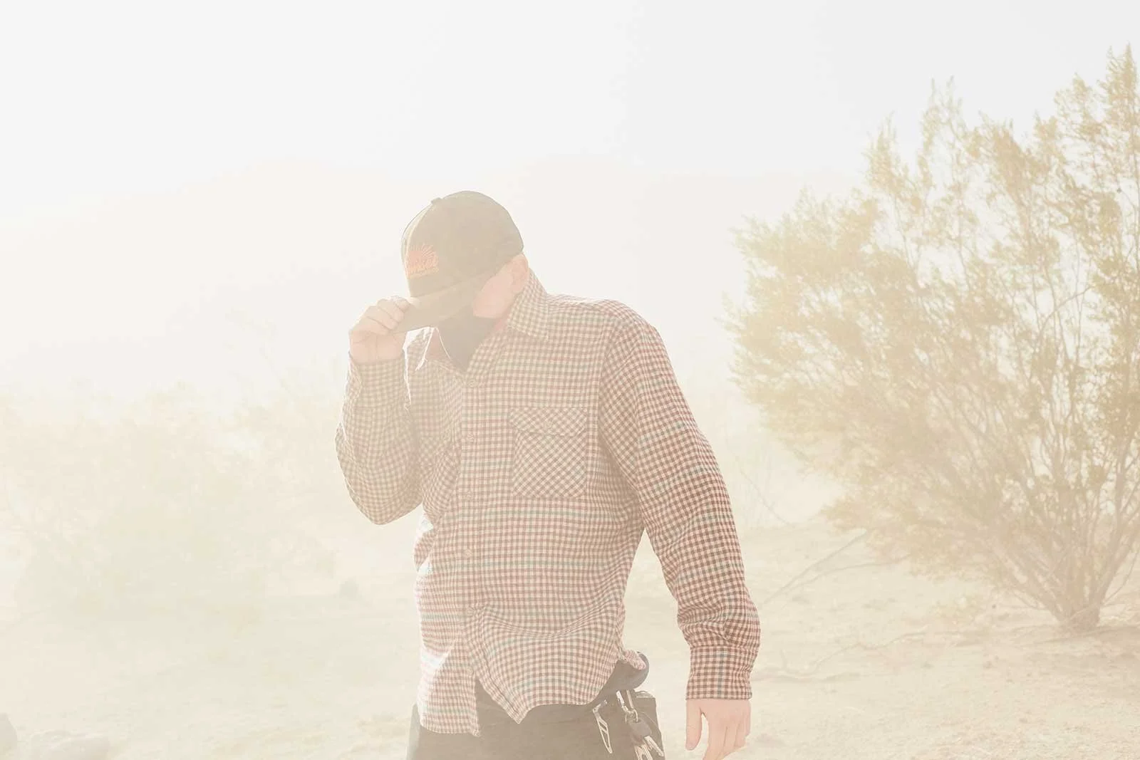A man wearing a checkered shirt, black hat, and gloves, standing outdoors in a foggy environment with trees in the background.