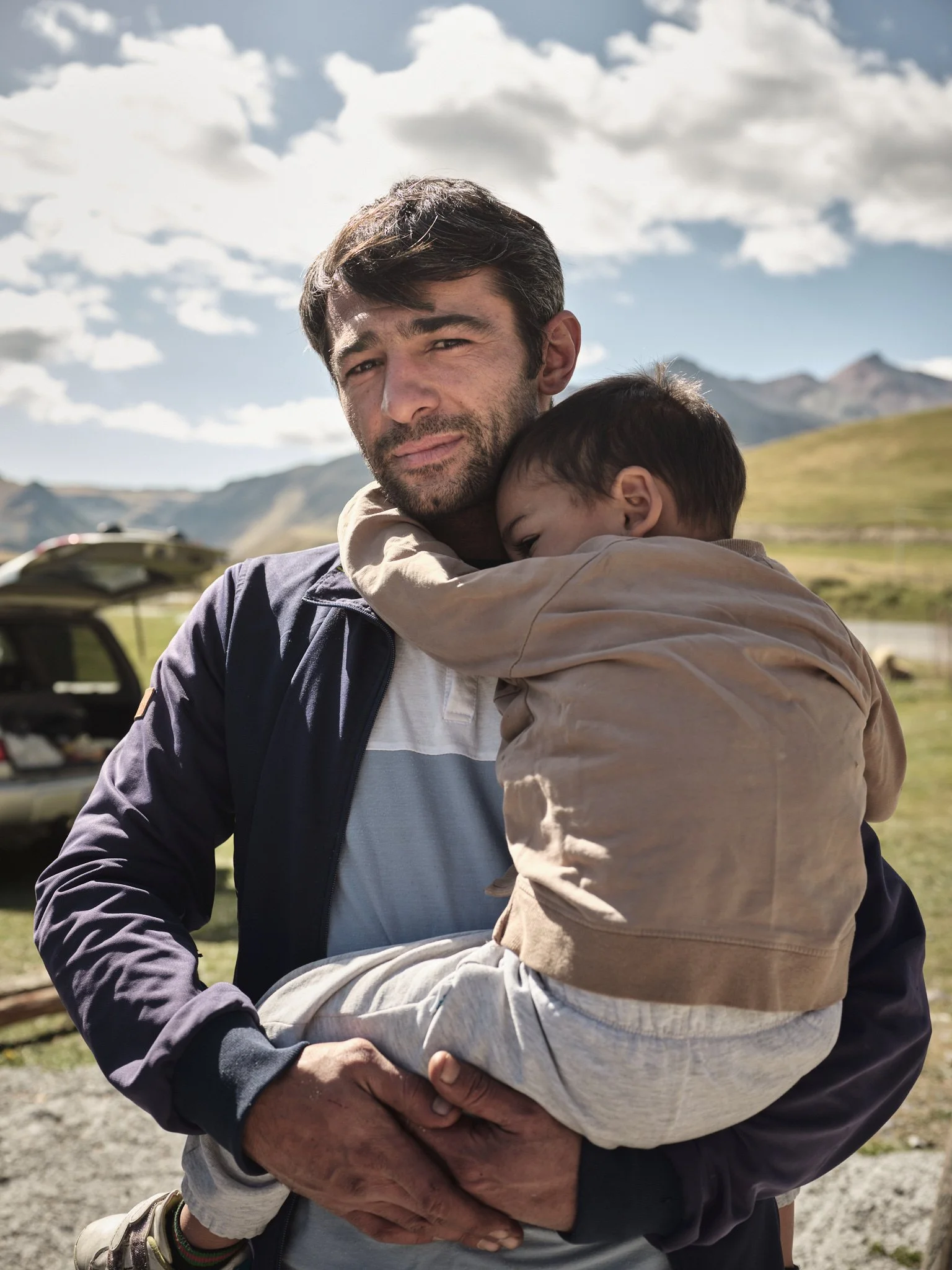 A man holding a young child in his arms outdoors with mountains and a partly cloudy sky in the background.