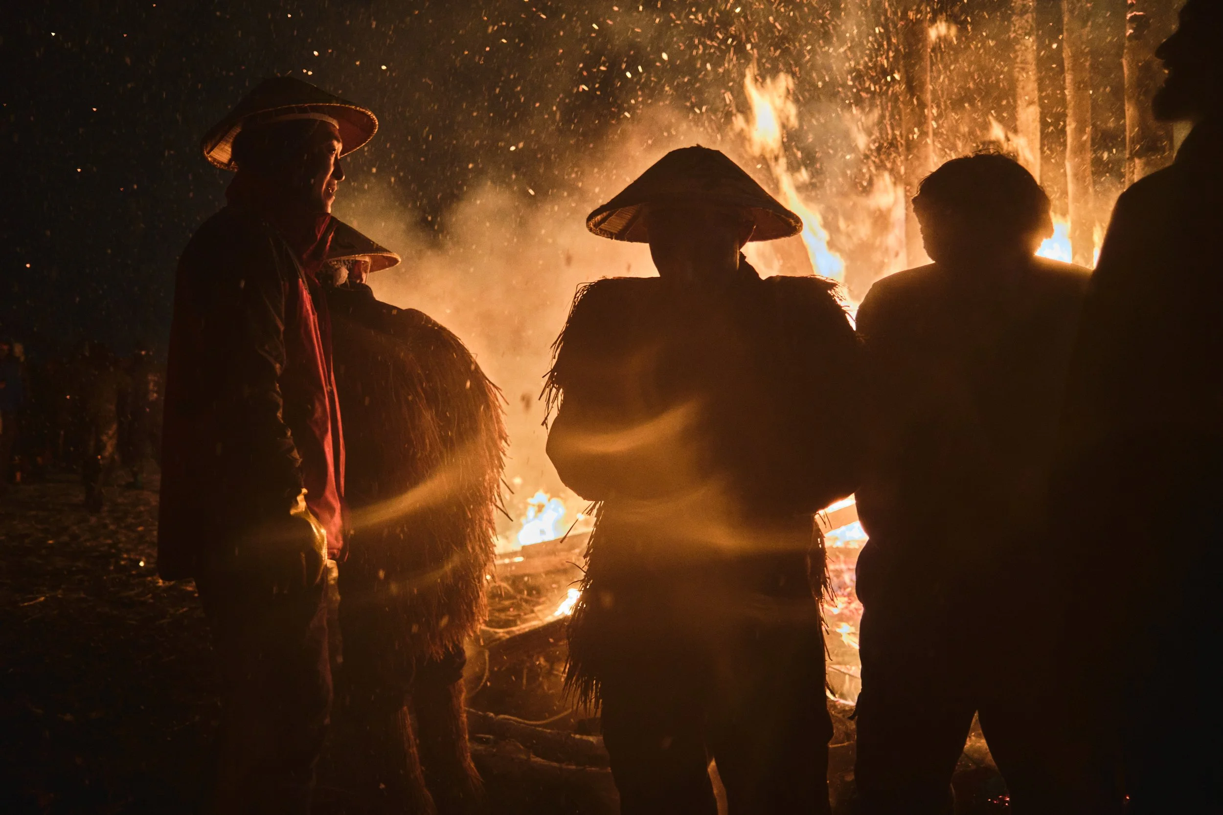 Silhouettes of five people wearing traditional hats standing around a large bonfire at night.