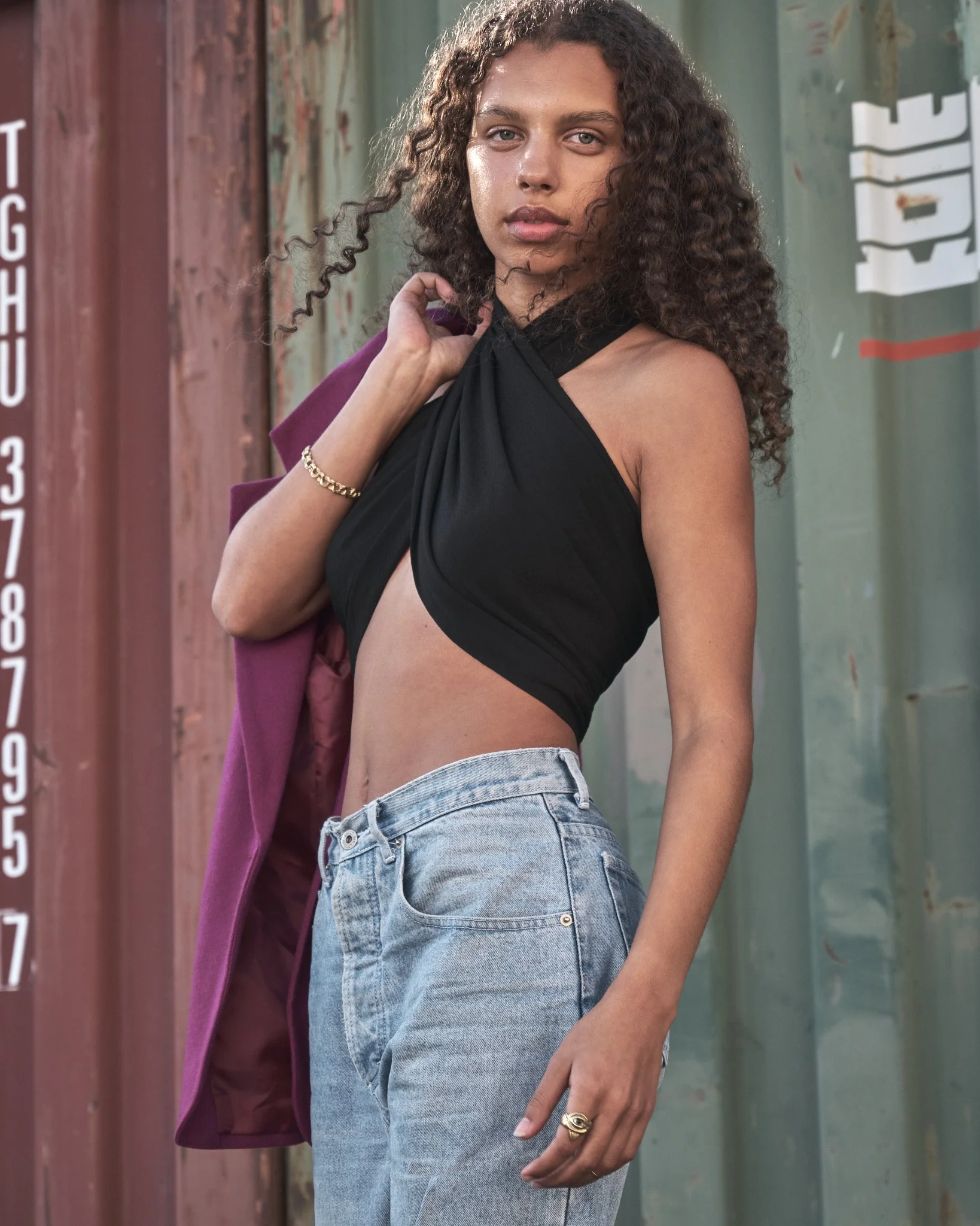 A young woman with curly hair standing outdoors near a weathered shipping container, wearing a black halter top, light blue jeans, and carrying a purple jacket over her shoulder.