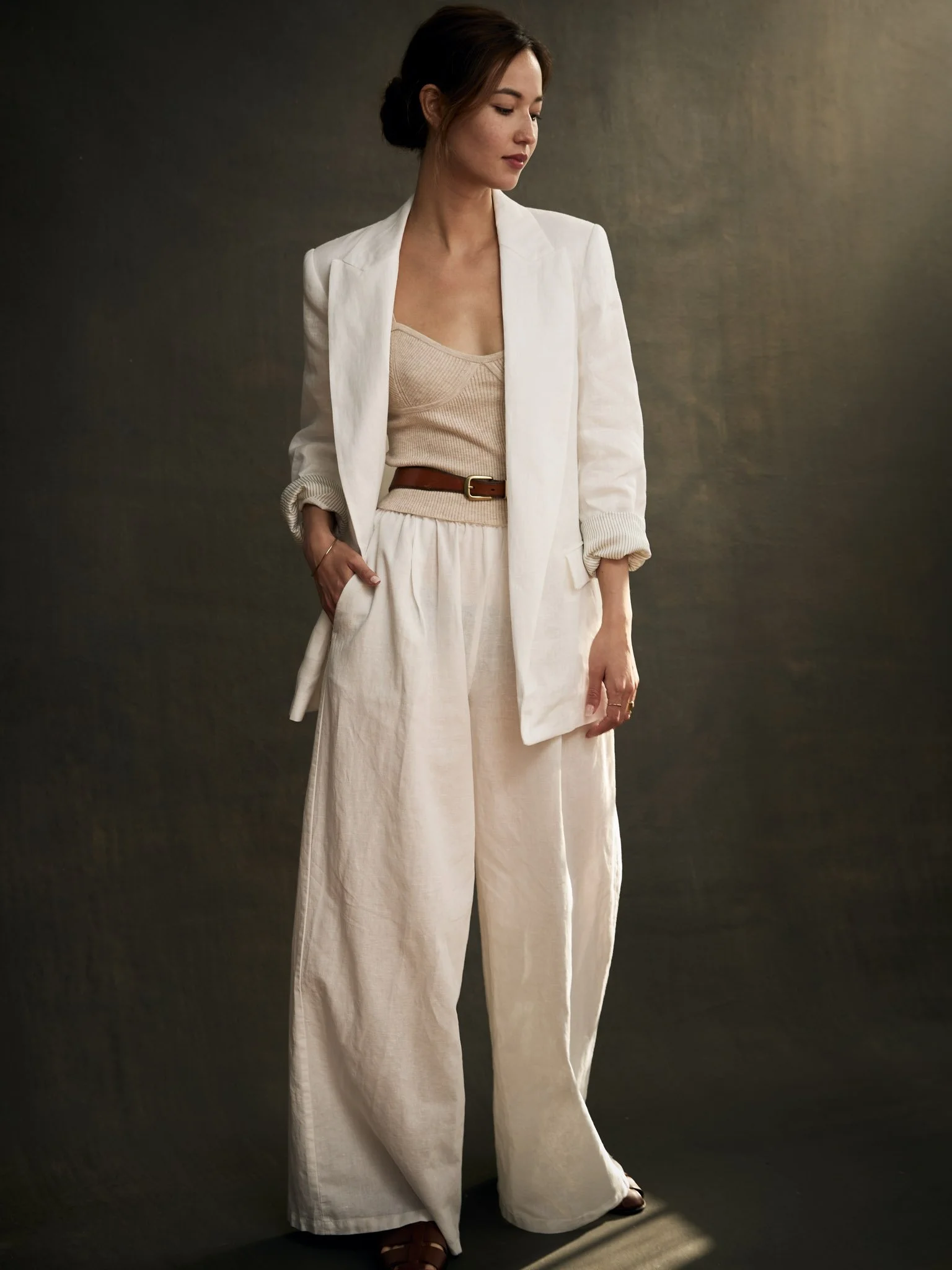 A woman in a cream-colored outfit with a blazer, wide-leg pants, and a sleeveless top, standing against a dark background.