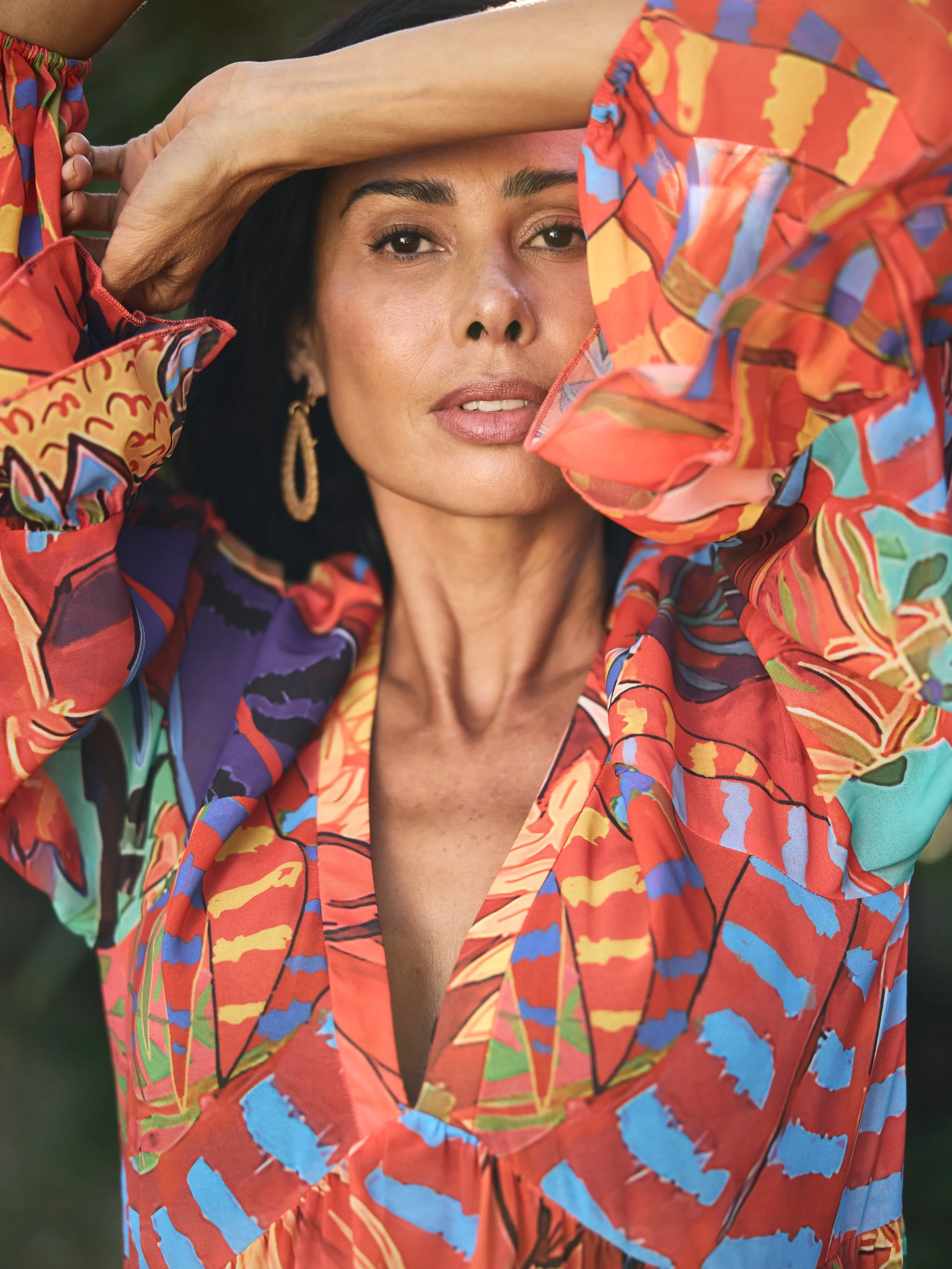 A woman with dark hair and tan skin, wearing gold hoop earrings, poses outdoors with her hands on her head. She is dressed in a colorful, abstract-patterned shirt with orange, red, blue, green, and yellow hues.
