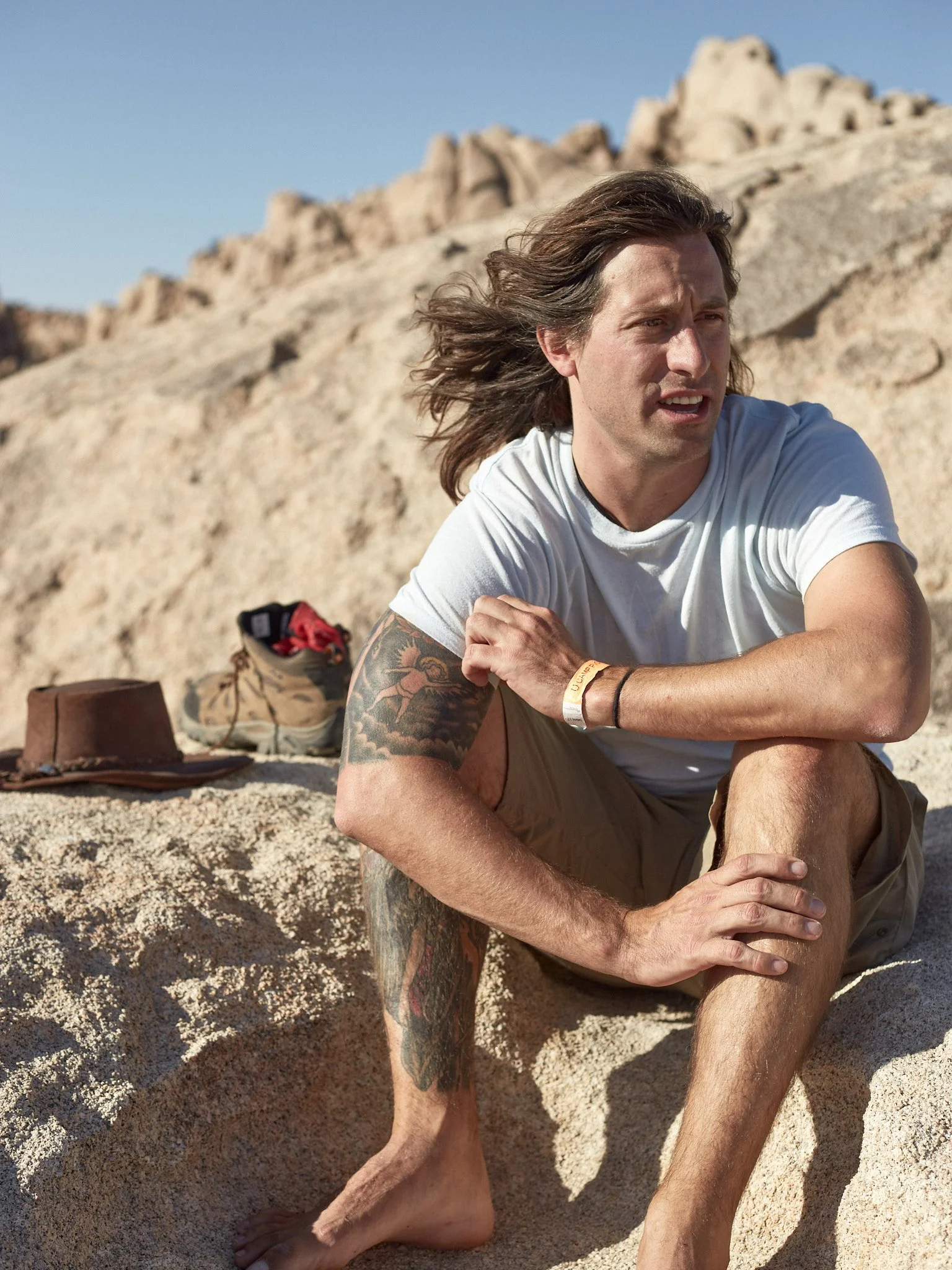 A man with shoulder-length brown hair, tattoos on his arms, wearing a white t-shirt and khaki shorts, sitting barefoot on a sandy rock in a desert-like environment with large rock formations in the background.