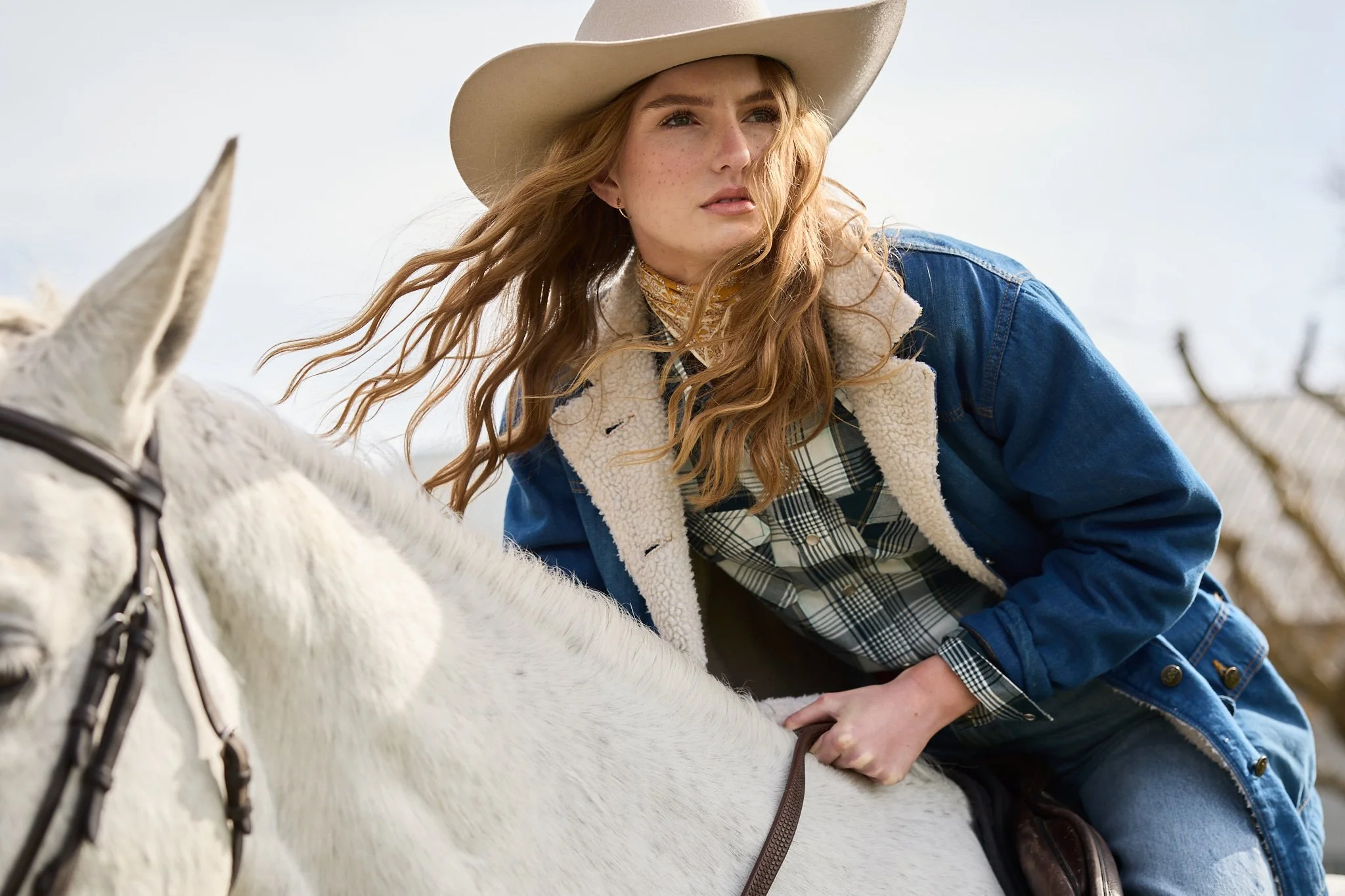 A red-haired woman wearing a cowboy hat and denim jacket riding a white horse outdoors.