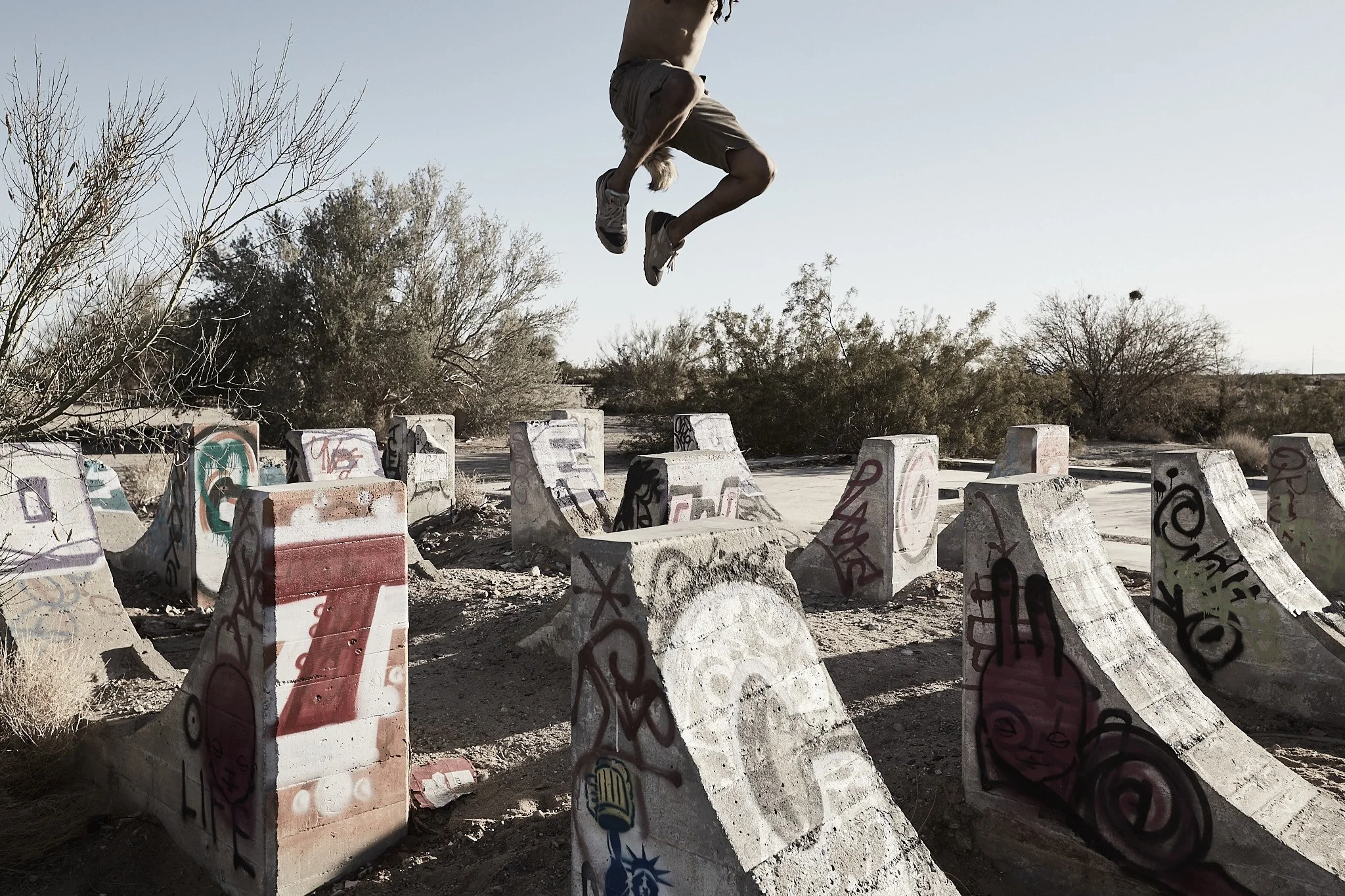 A person performing a jump at an outdoor skate park with graffiti-covered concrete ramps and bowls, and dry desert vegetation in the background.