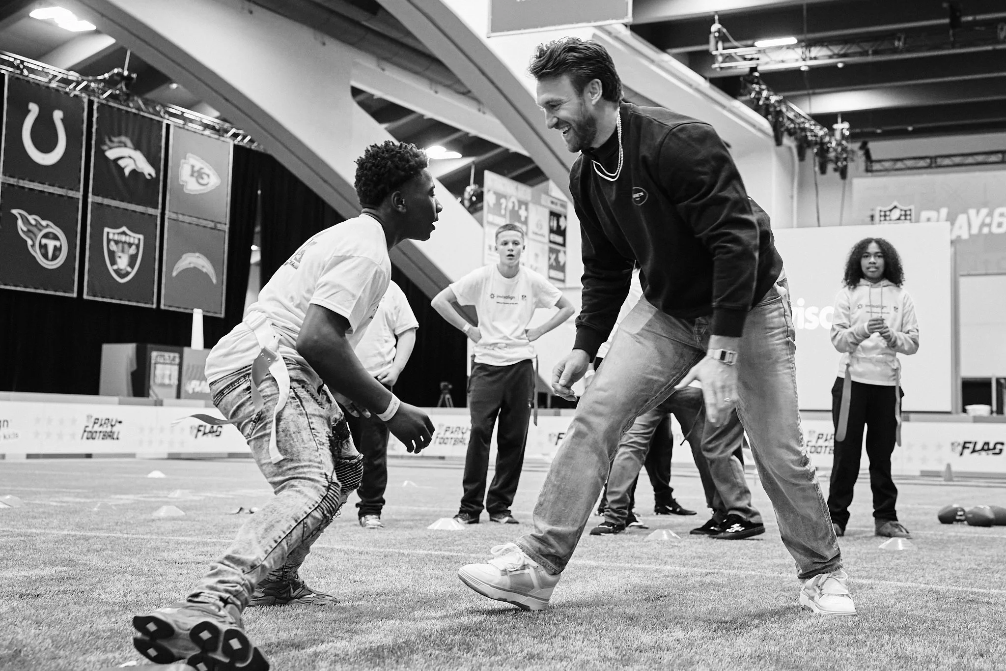 NFL fullback Kyle Juszczyk plays flag football with a young special olympics player engaged in a training drill on the field, with other children observing in the background, inside an indoor sports facility. Photographed for Invisalign.