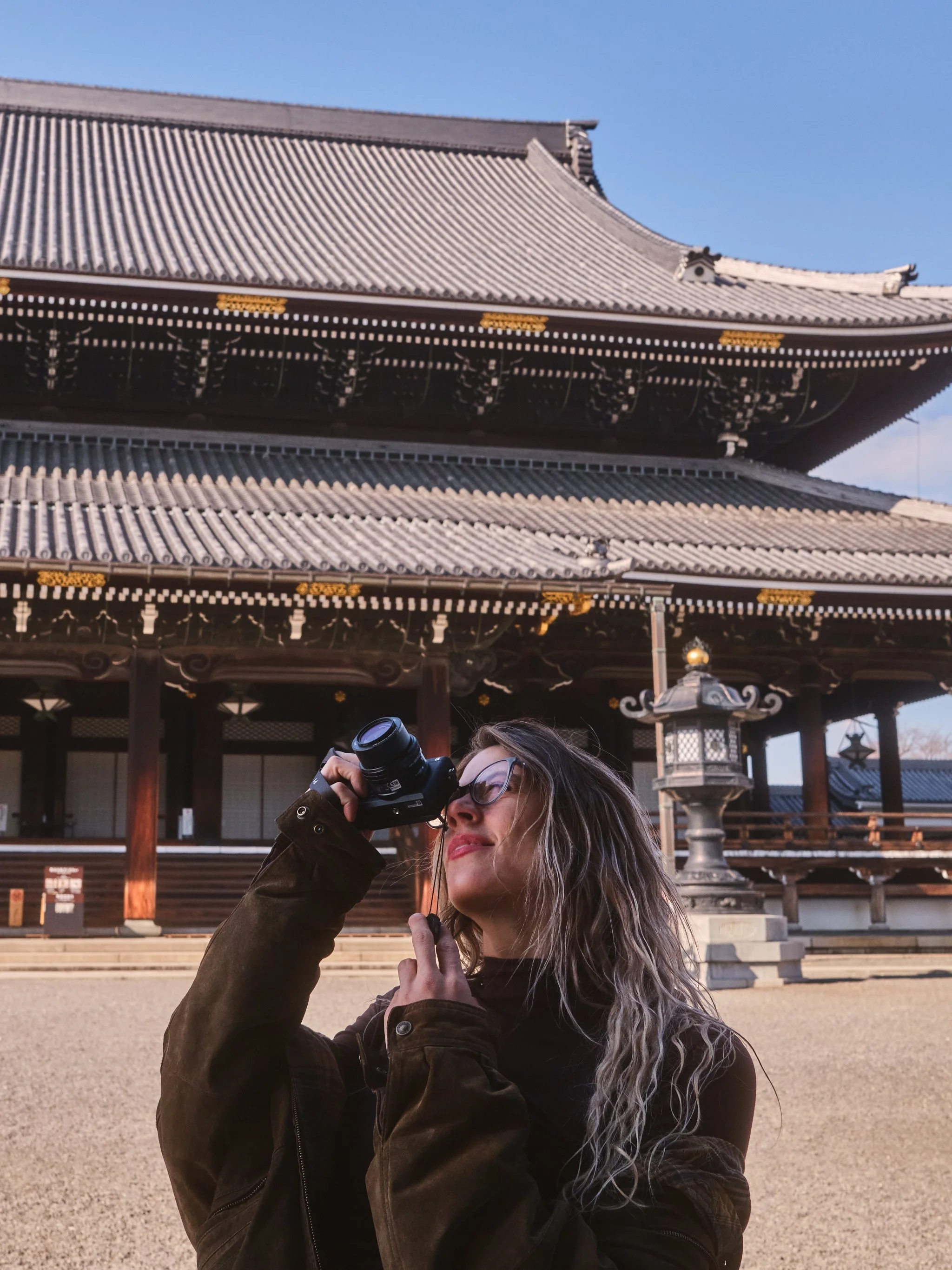 A woman with wavy hair and glasses is taking a photograph with a camera in front of a traditional Japanese temple.
