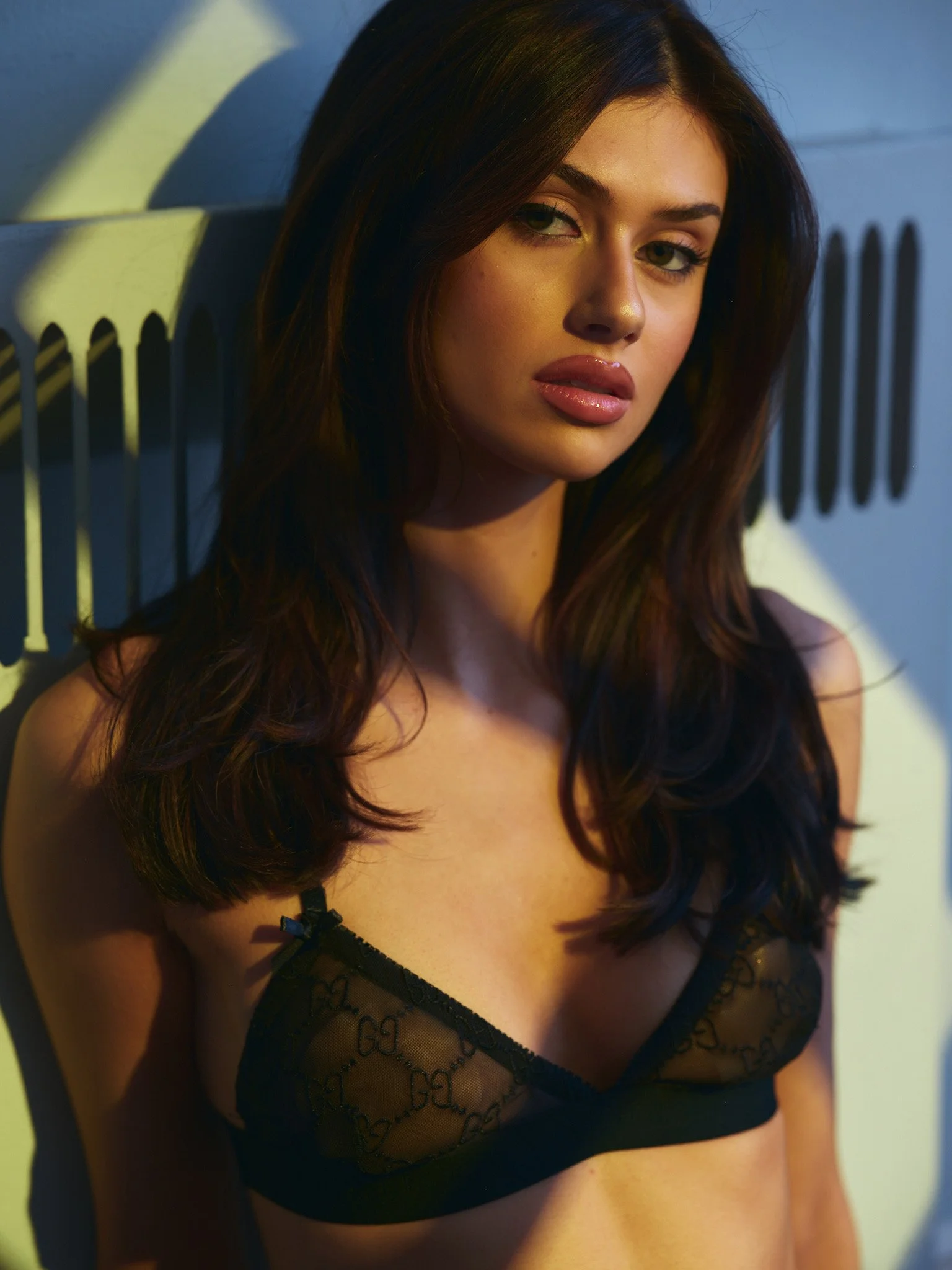 Lingerie photoshoot with dynamic lighting of a model with long, dark hair wearing a black lace bra, leaning against a painted wall, looking at the camera with a neutral expression.
