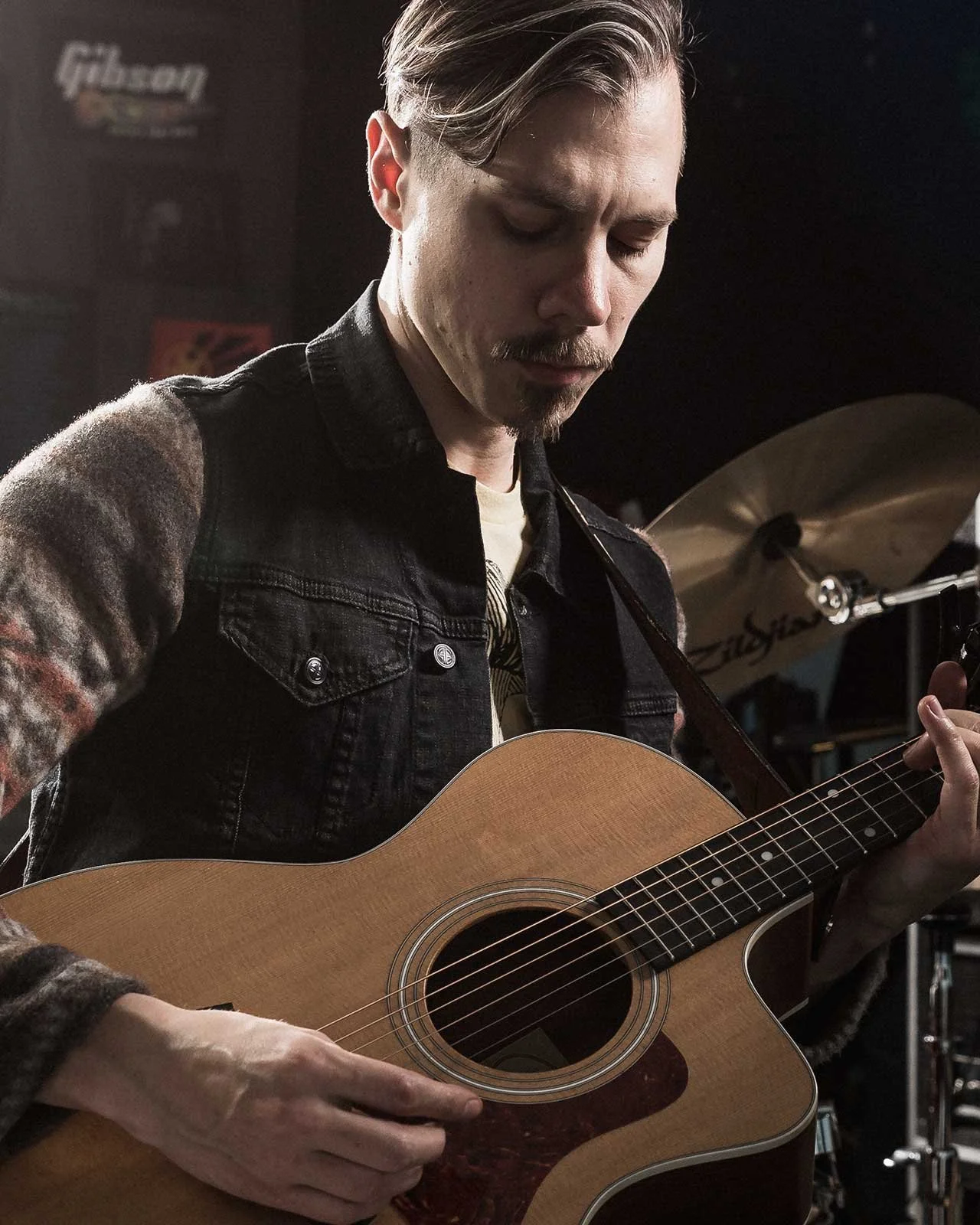 A man with light skin, styled hair, and a mustache playing an acoustic guitar in a dimly lit room, with a drum set visible in the background.