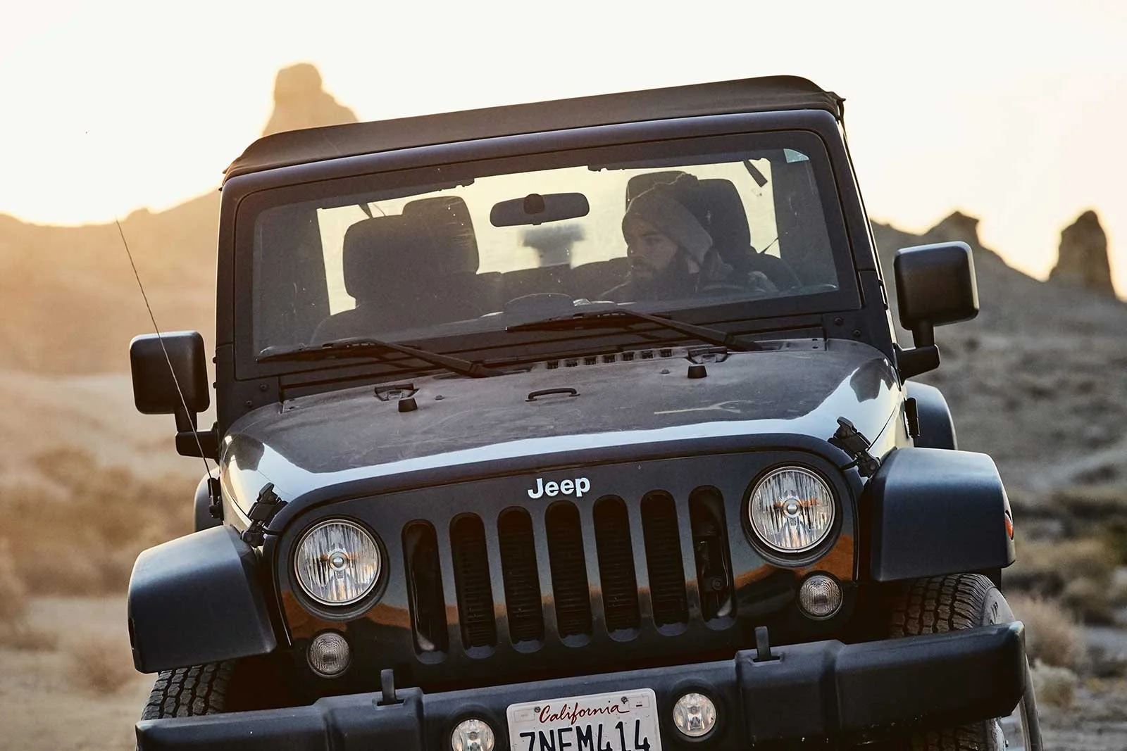 A black Jeep Wrangler with a California license plate driving on a rocky trail in a desert landscape at sunset, with a man in the driver seat wearing a knit cap.
