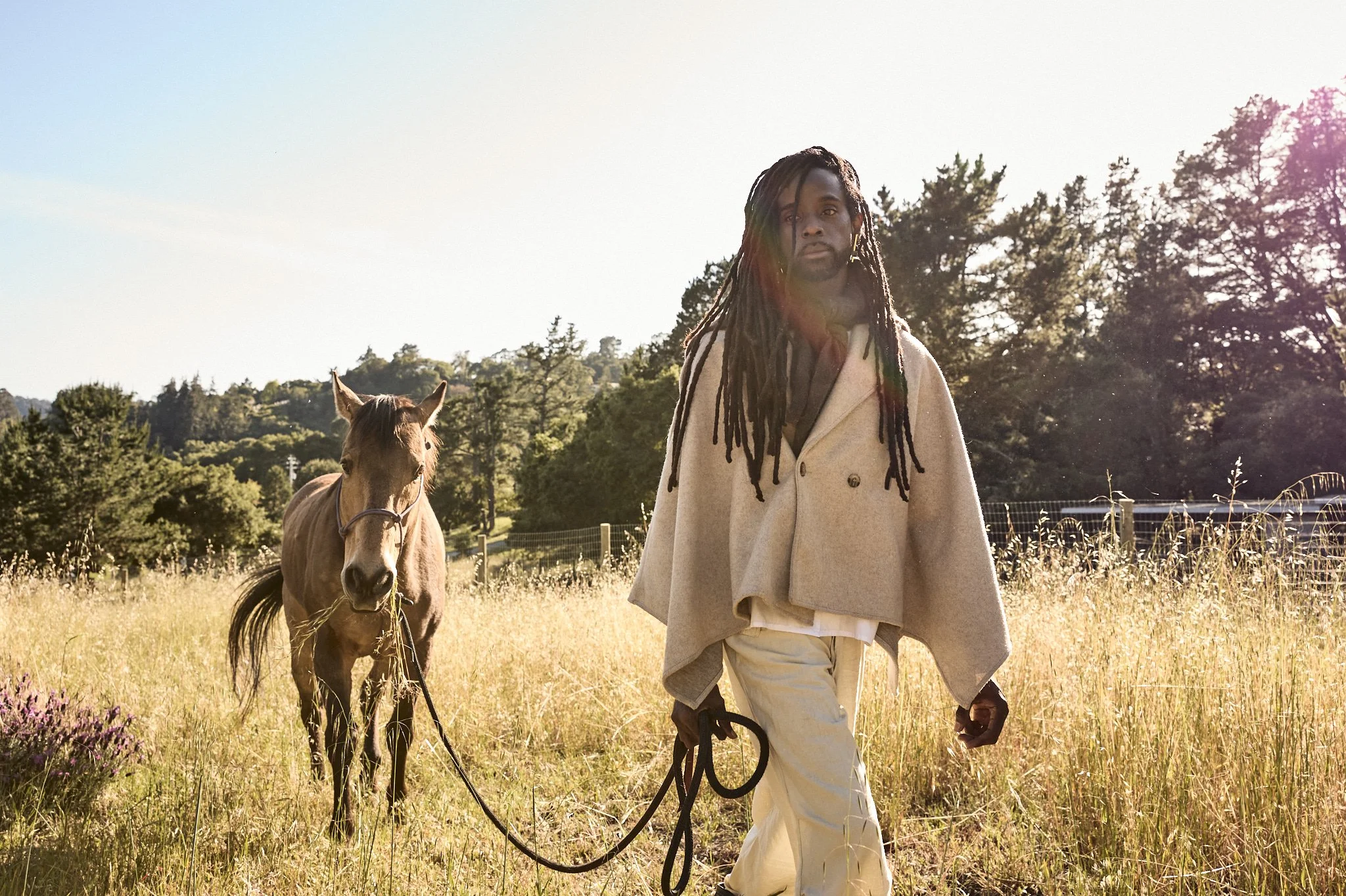 A man with long dreadlocks walking a horse in a grassy field during sunset.