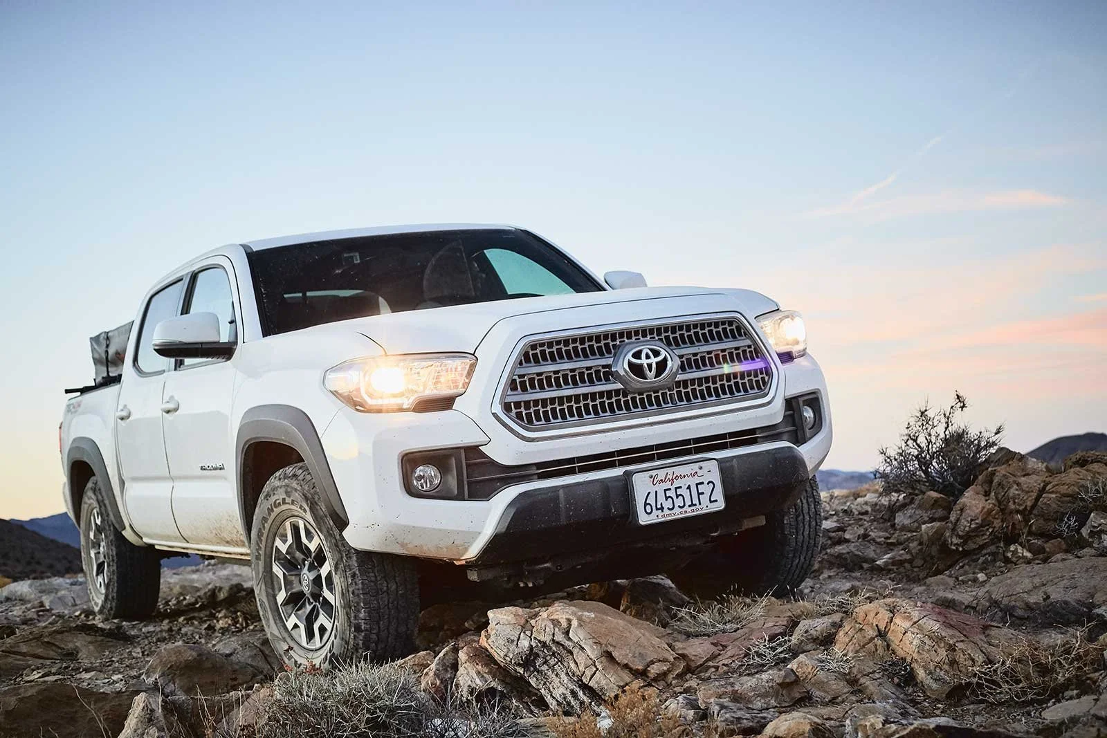 A white Toyota pickup truck driving on rocky terrain at sunset, with desert landscape and mountains in the background.