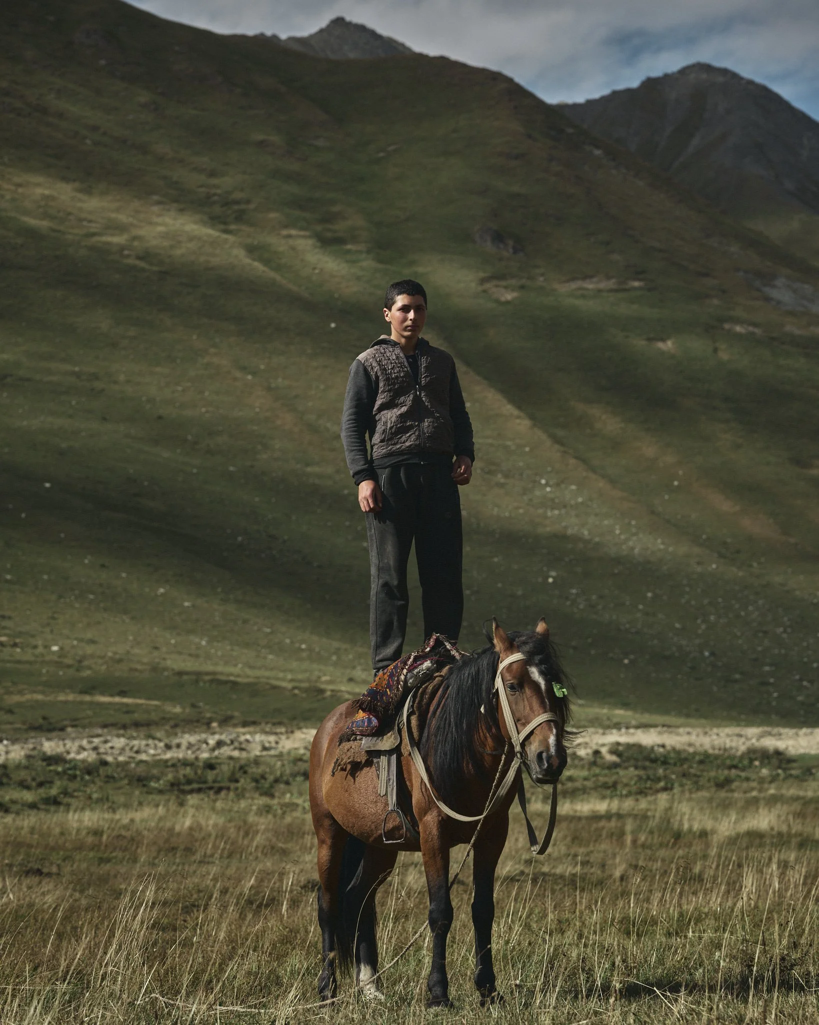 Young boy standing on a horse in a grassy field with mountains in the background.