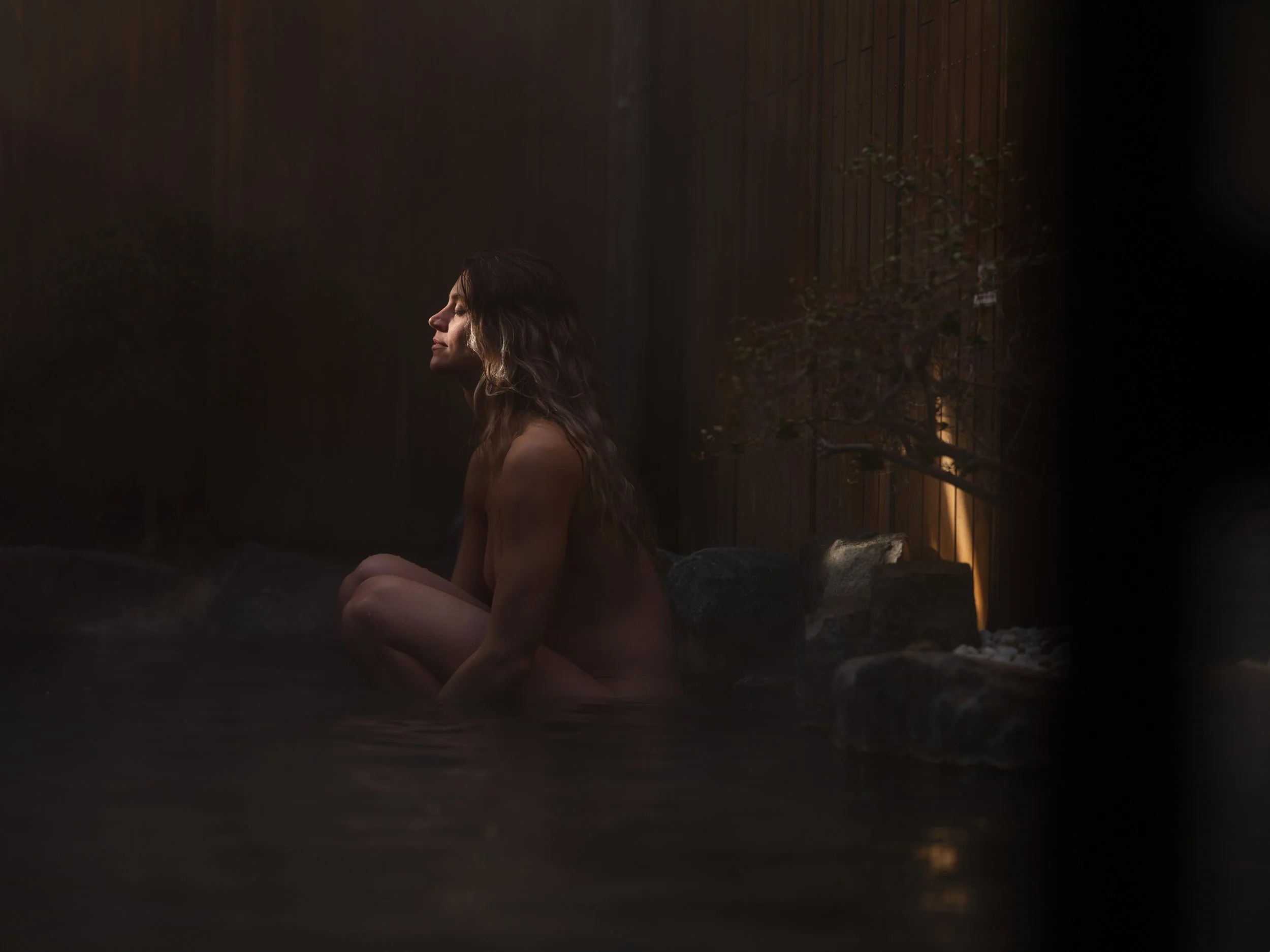 A woman with long, wavy hair sitting in a hot spring or bath at dusk or night, with a wooden fence and a small tree in the background and soft warm lighting.