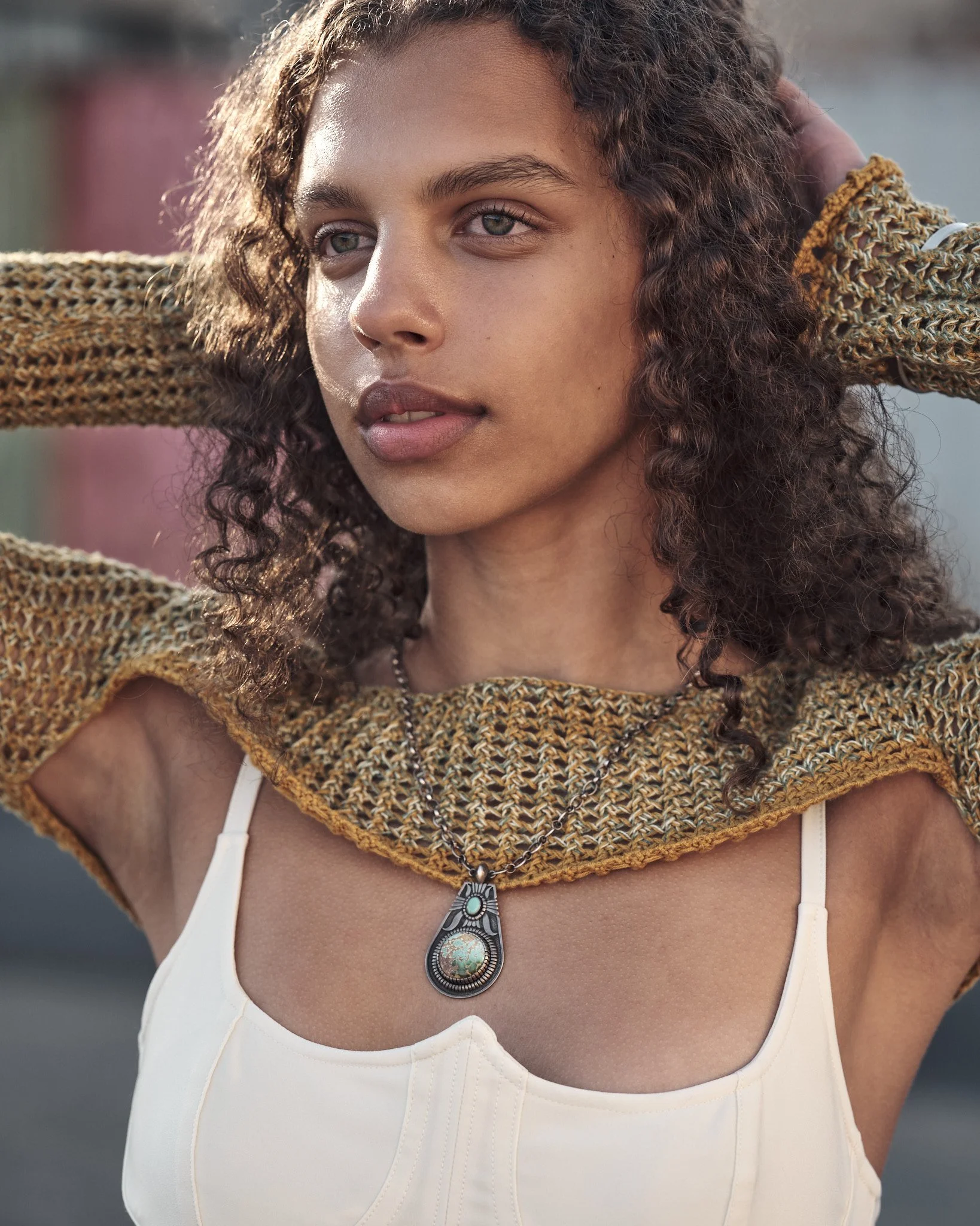 Photo of a young woman with curly hair wearing a patterned shawl and a white top, displaying a necklace with a large pendant, outdoors in natural light.