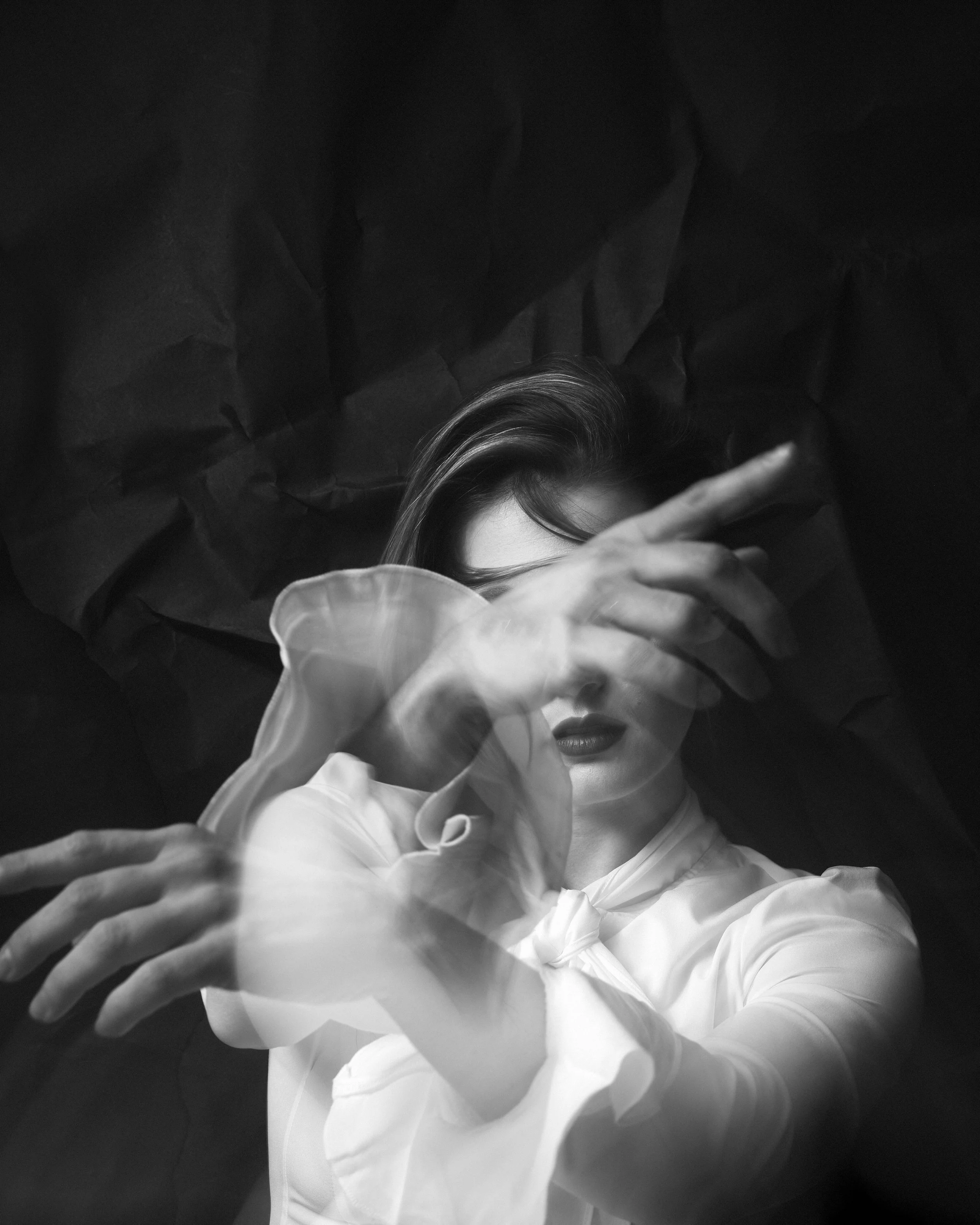 Black and white photograph of a woman lying on a dark surface with her face partially obscured by her raised arm and hand, wearing a light-colored blouse and lipstick.