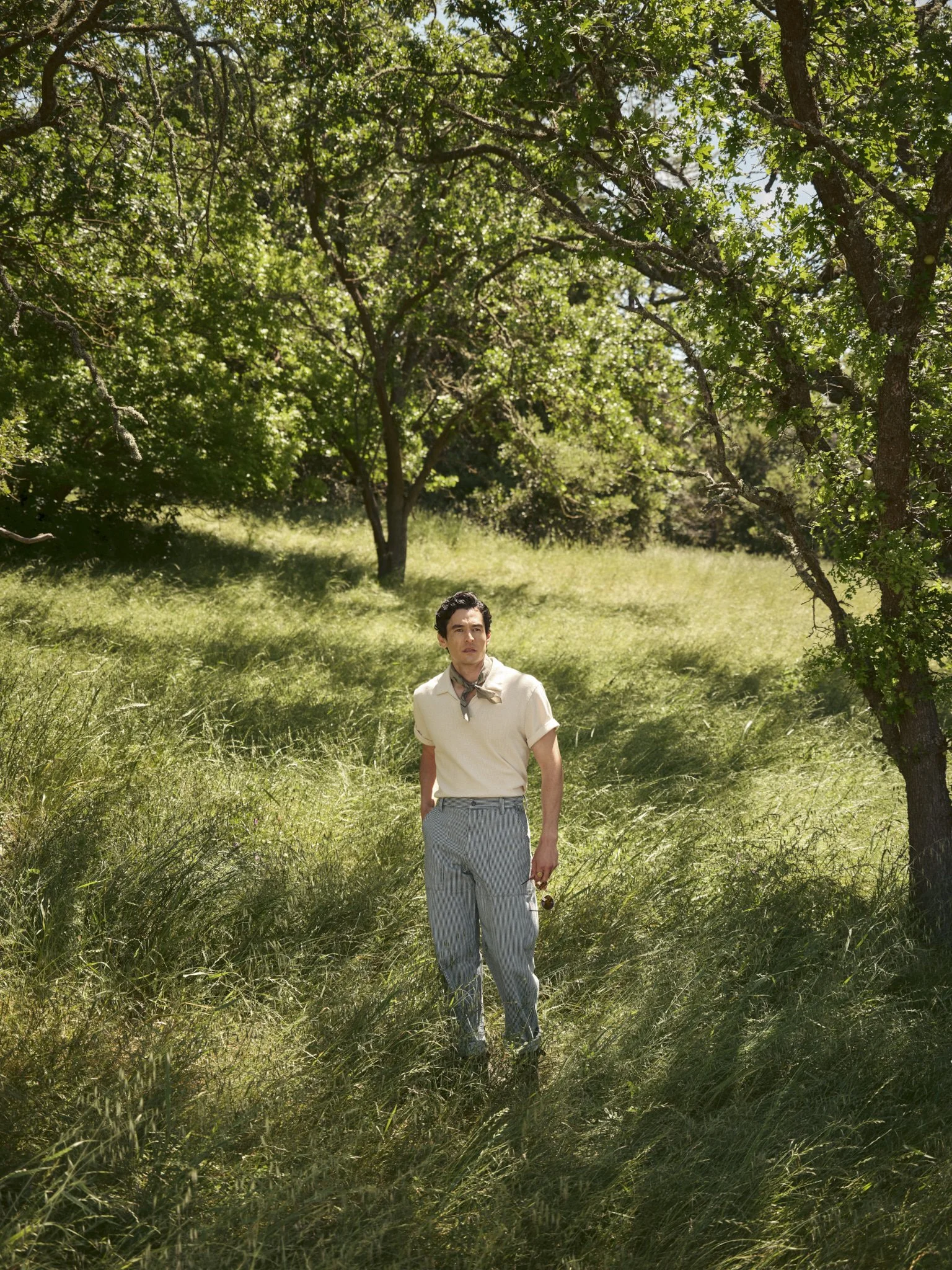 A man stands in a grassy field under the shade of trees, dressed in vintage clothing, holding sunglasses in his right hand, with a thoughtful expression.