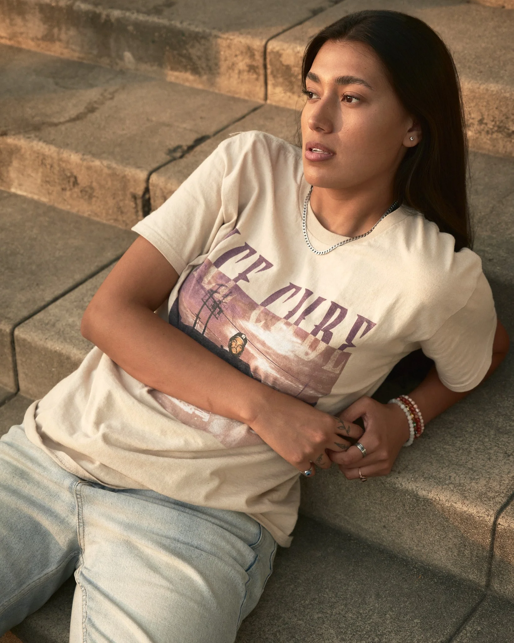 A young woman with long dark hair and tattoos on her hand, lying on concrete steps outdoors at sunset, wearing a graphic t-shirt and bracelets.