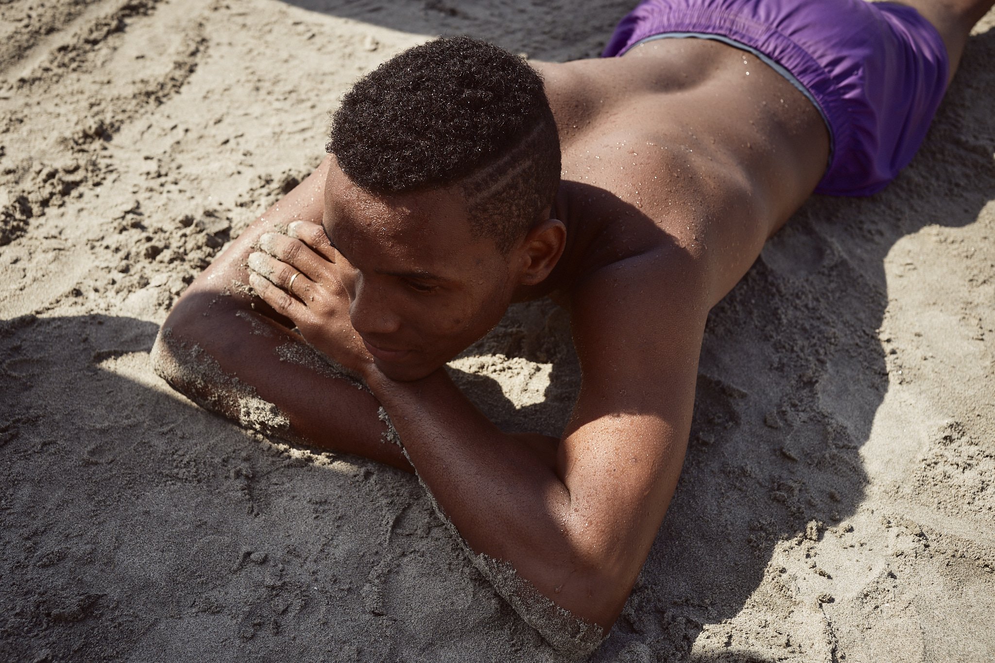 A young man lying on sandy beach, resting his head on his left arm and gazing downward.