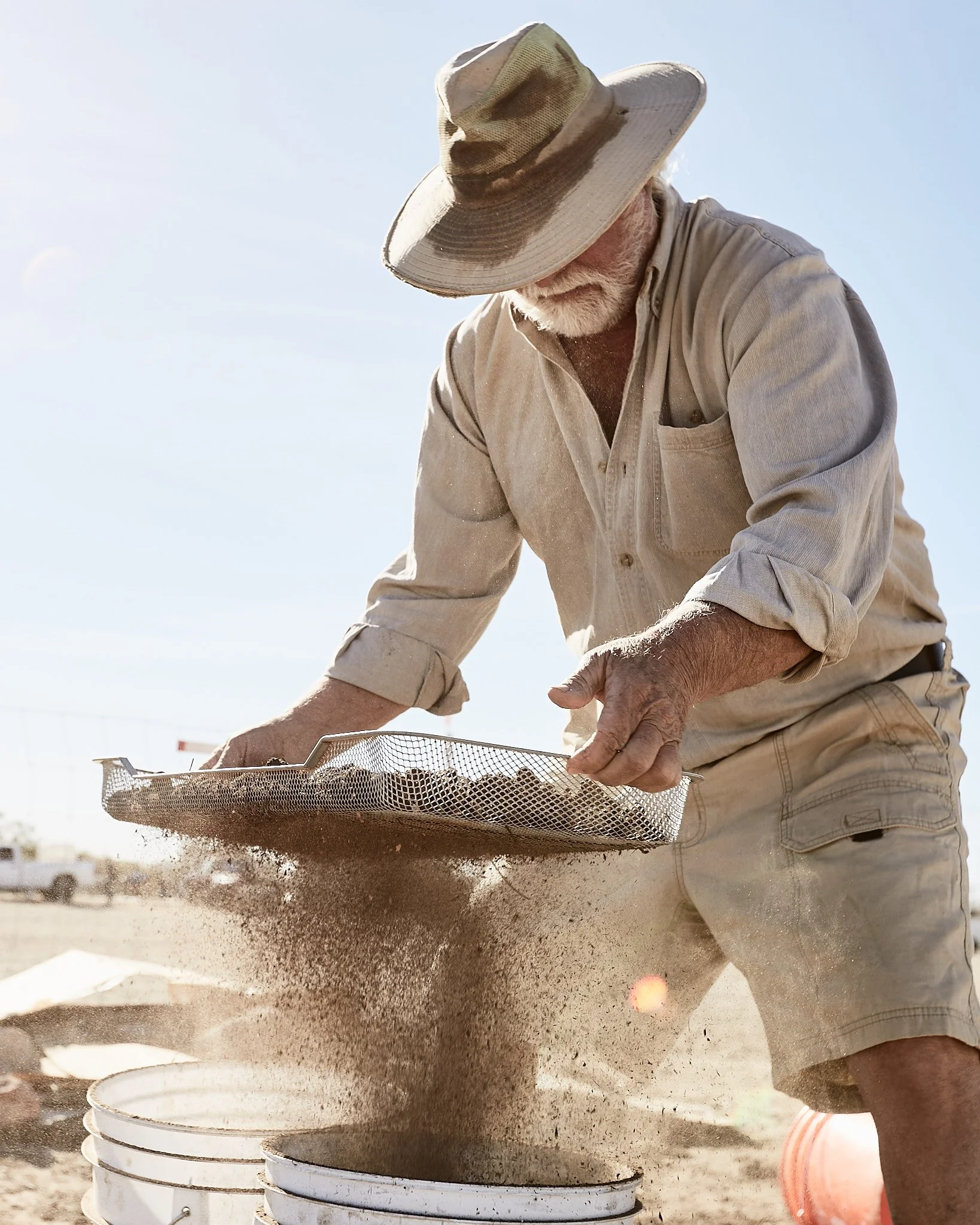 An elderly man wearing a wide-brimmed hat, a beige shirt with rolling sleeves, and khaki shorts sifting soil or sand through a metal screen into a white bucket outdoors on a sunny day.