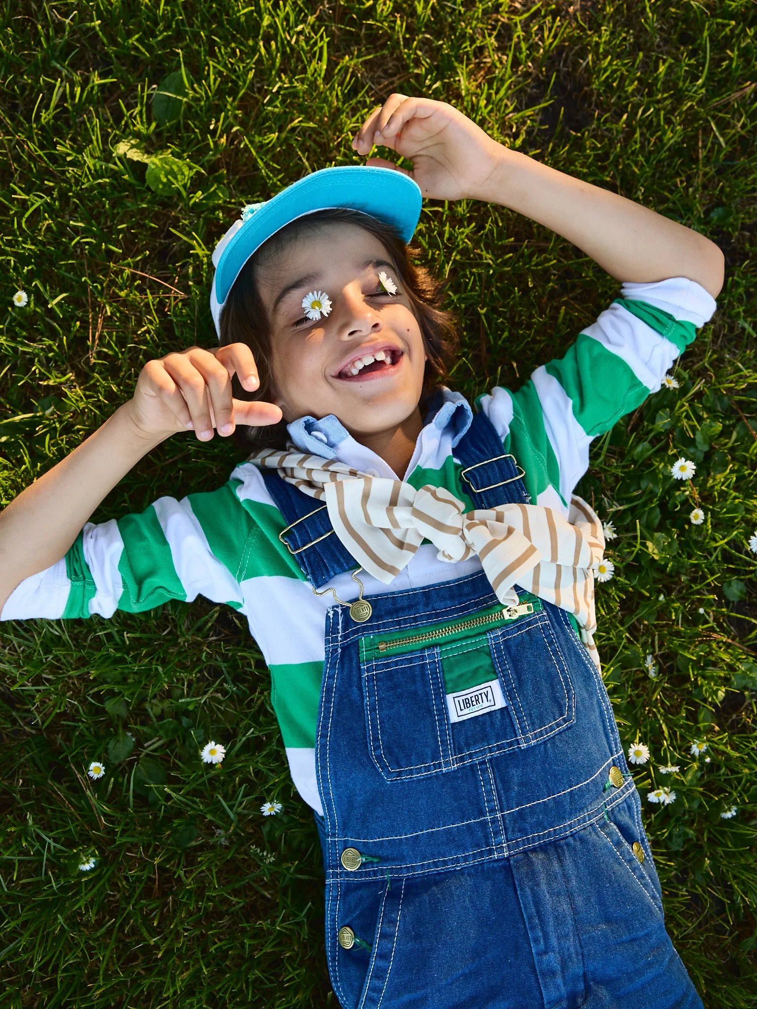 Young girl lying on grass with daisies, smiling, wearing a blue cap, striped shirt, denim overalls, and a striped scarf, with daisies on her face.