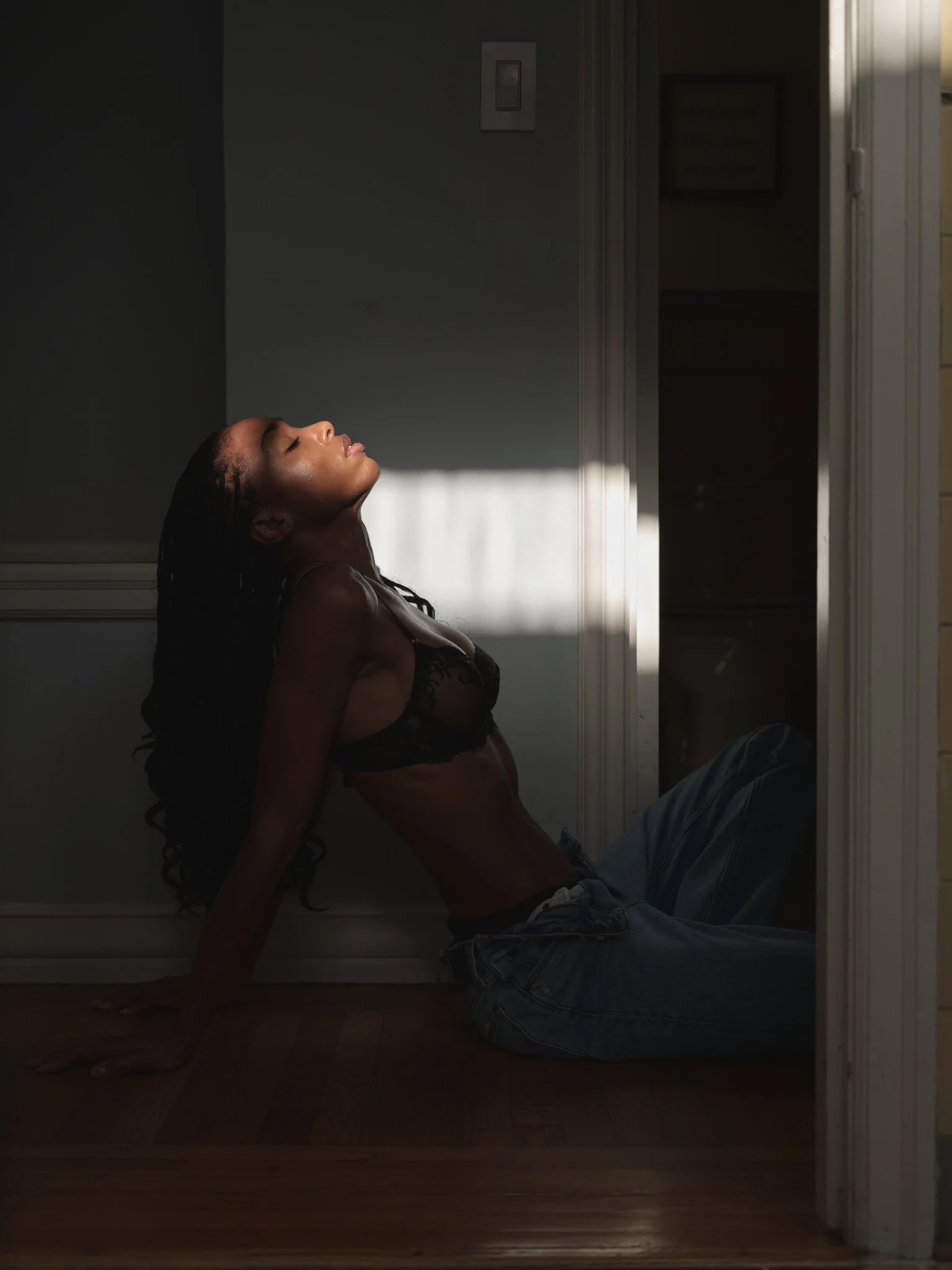 A woman with dark, curly hair reclining on the floor in a doorway, wearing a black lace top and blue jeans, with sunlight creating shadowy lighting.