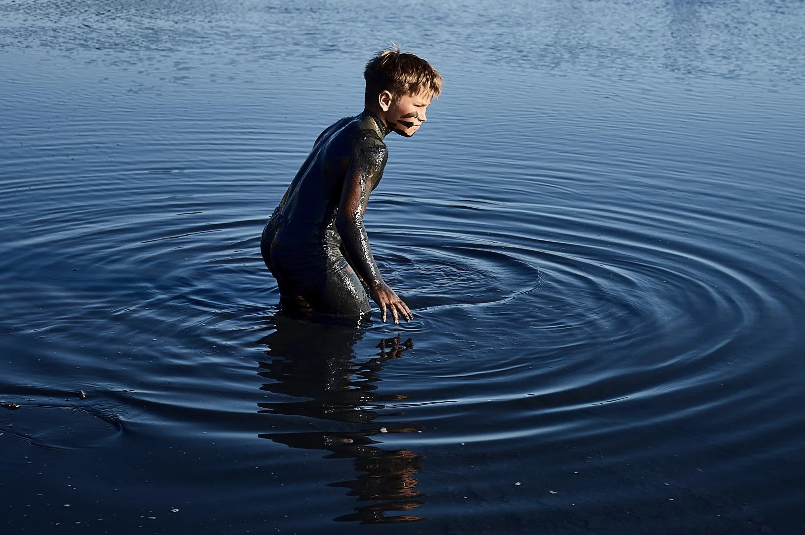 Child standing in a body of water with dark water, wearing a black wetsuit, with mud or black paint on their face and hands, immersed up to their waist, facing to the left.